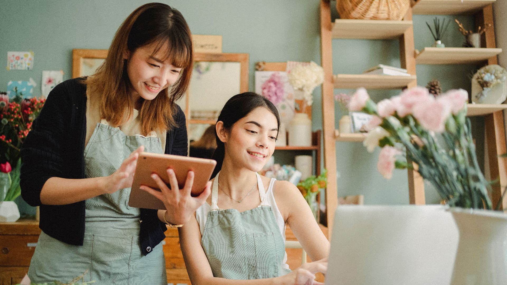Cheerful multiethnic female coworkers wearing aprons at table with flowers browsing on tablet and netbook while working online in floristry studio