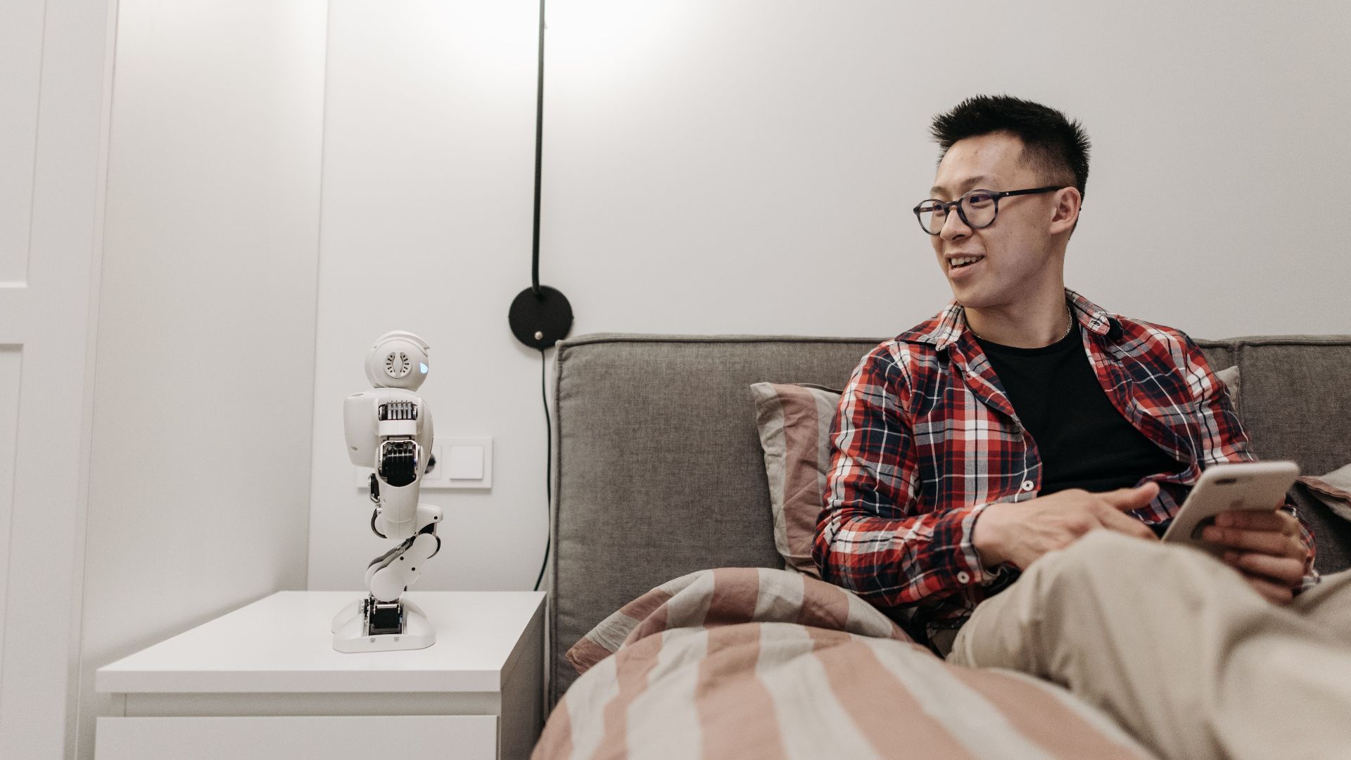 Man sitting on bed using mobile phone, interacting with small robot on bedside table.