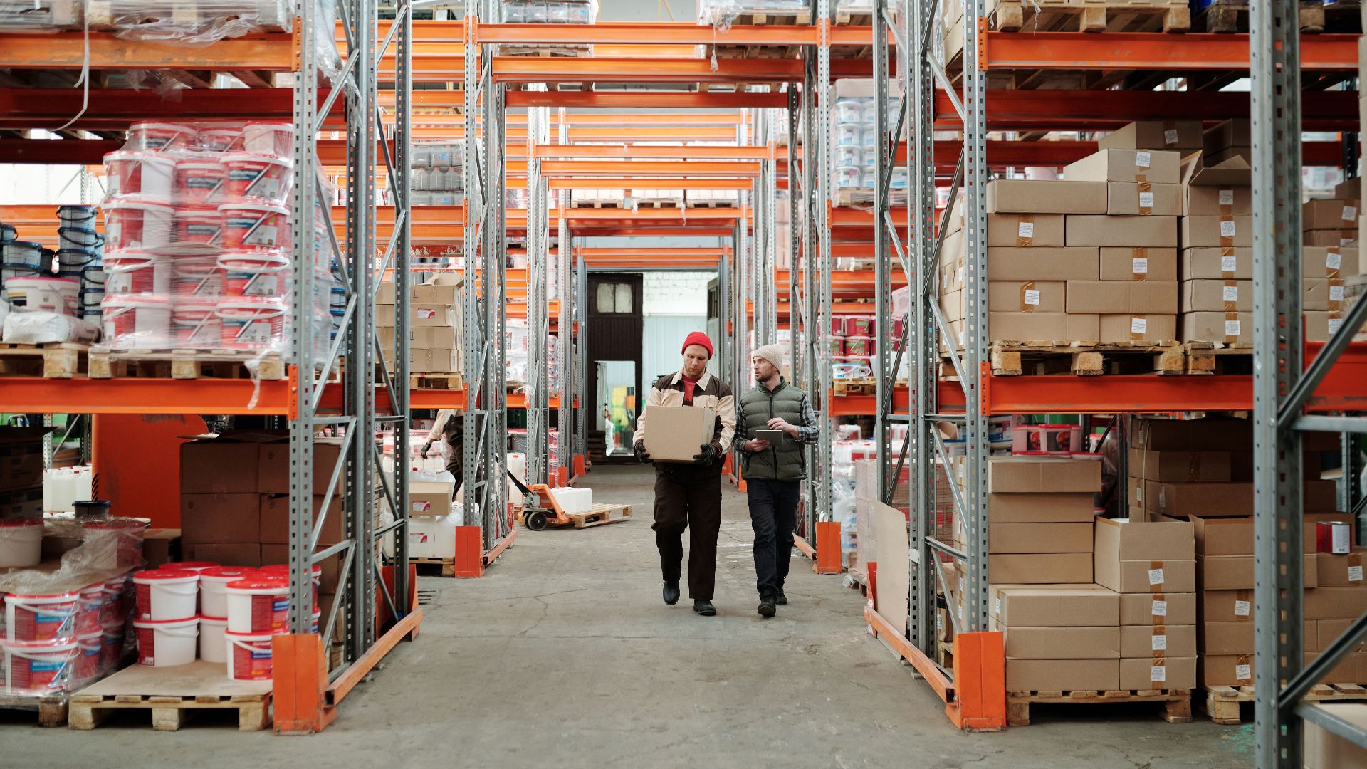 Two workers handle a package in a spacious warehouse surrounded by shelves stocked with boxes and products.