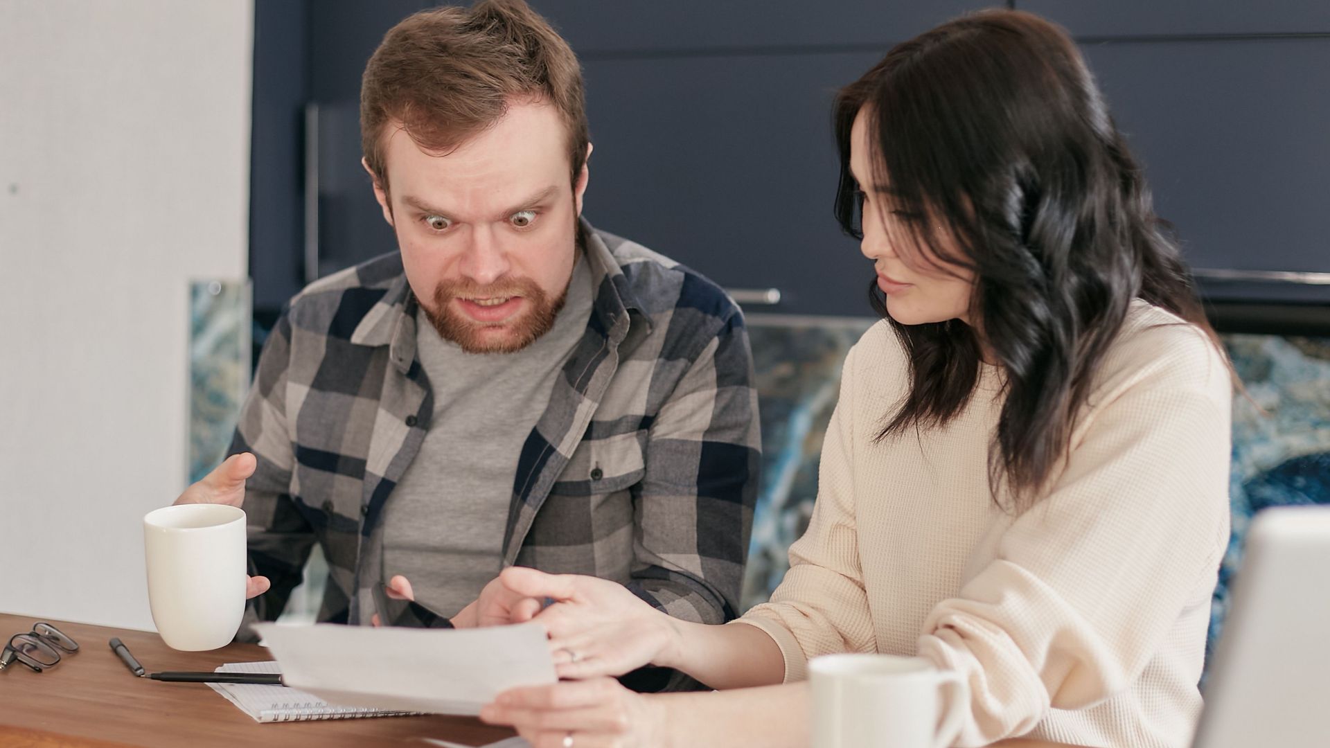 A couple shows astonishment as they review documents together at a wooden desk indoors.