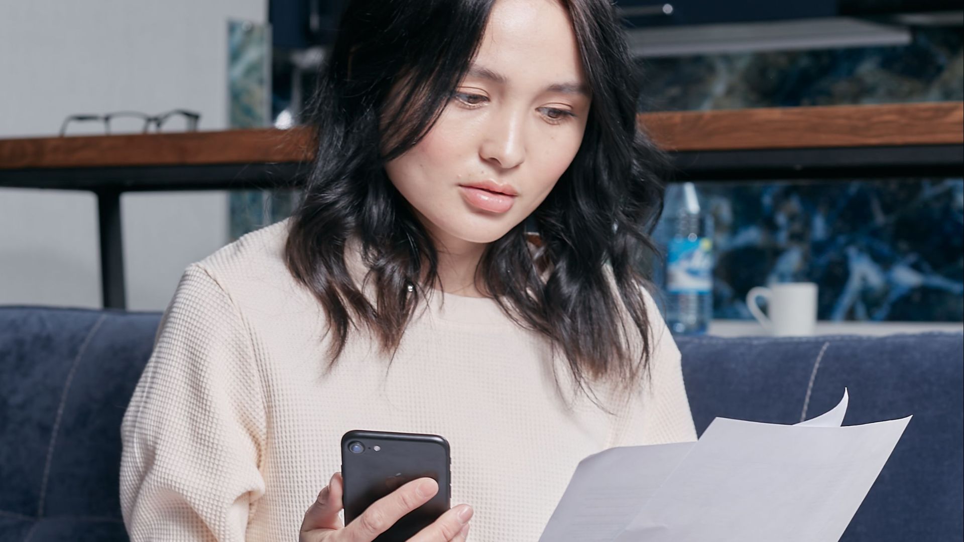 Young woman checking financial documents while sitting on a sofa with a smartphone and laptop.