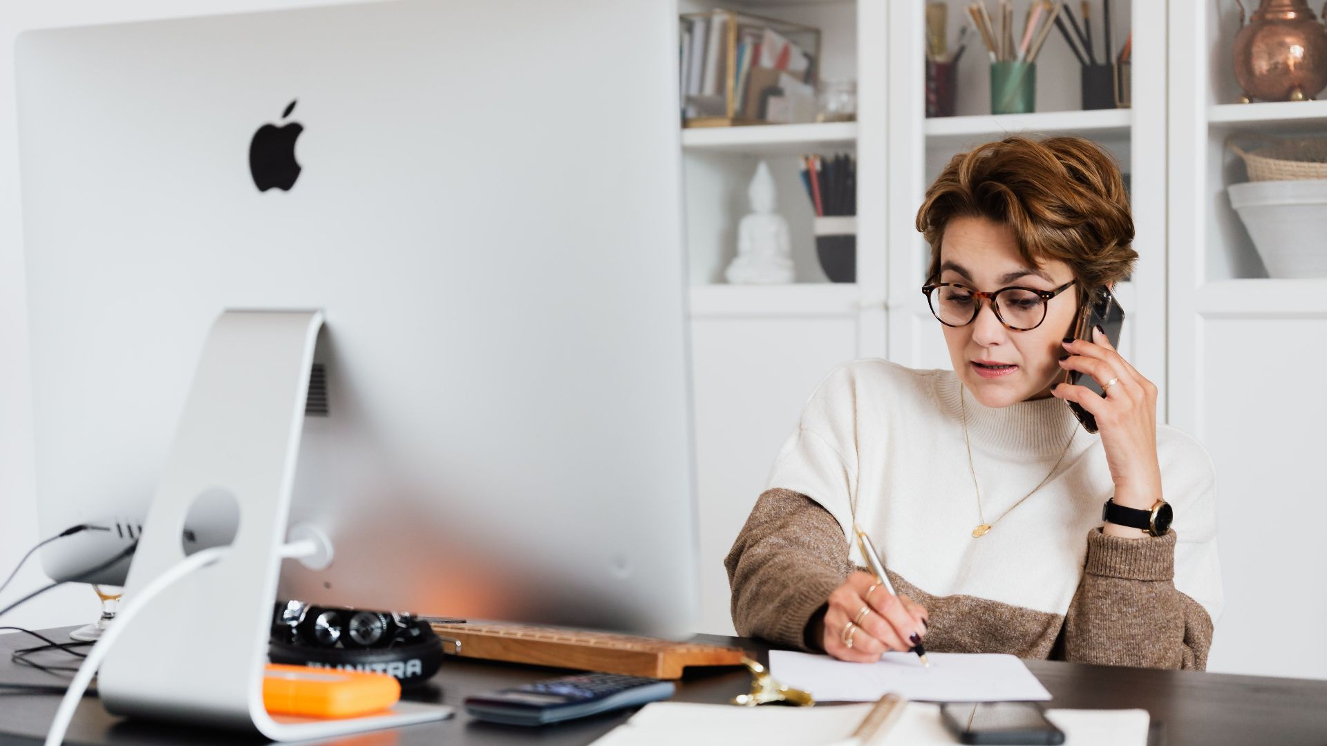 Busy woman in glasses communicating on mobile and writing down information while sitting at desk with modern monoblock computer in cozy study