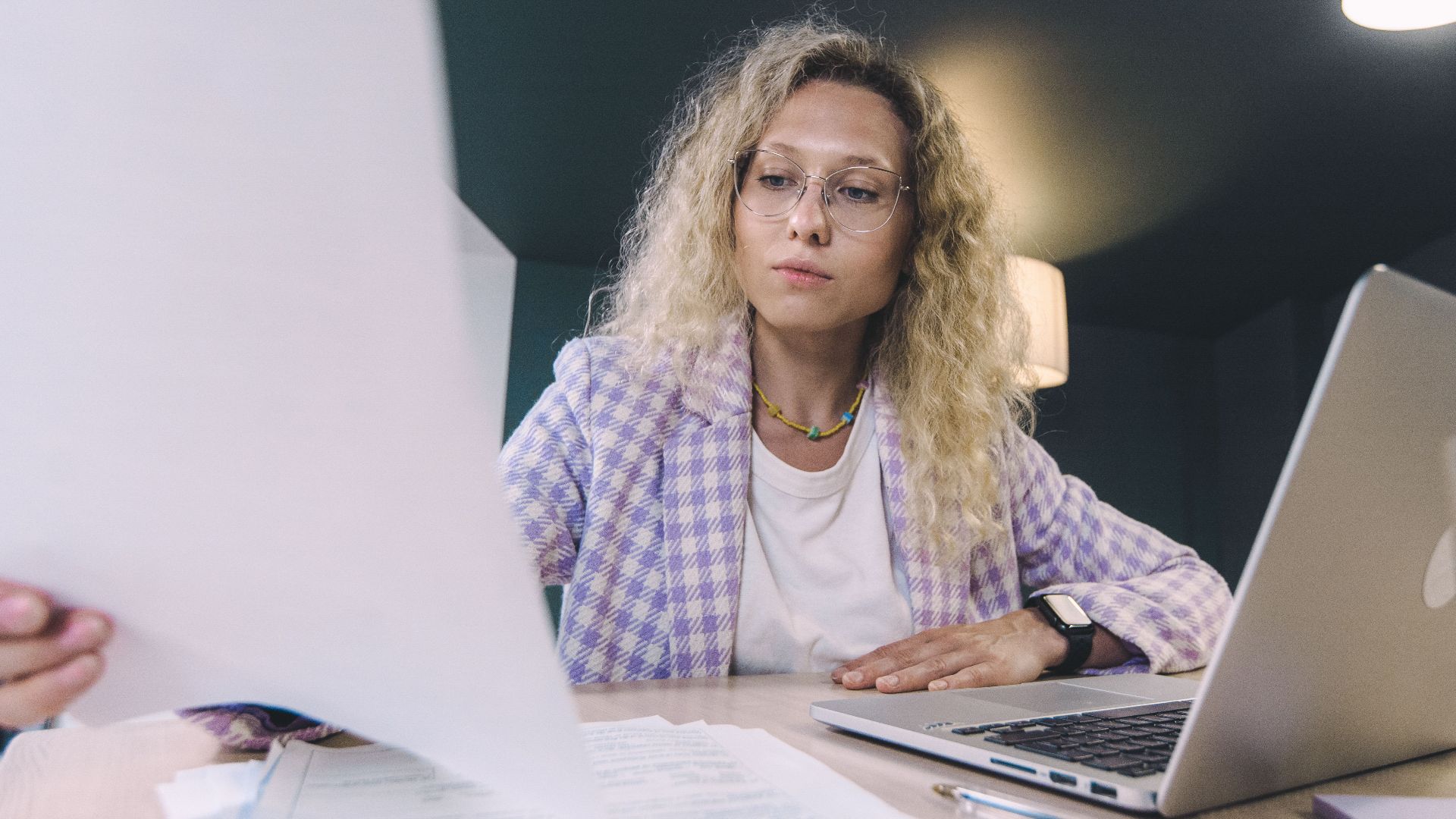Woman in an office environment reviewing documents with focus, surrounded by technology.