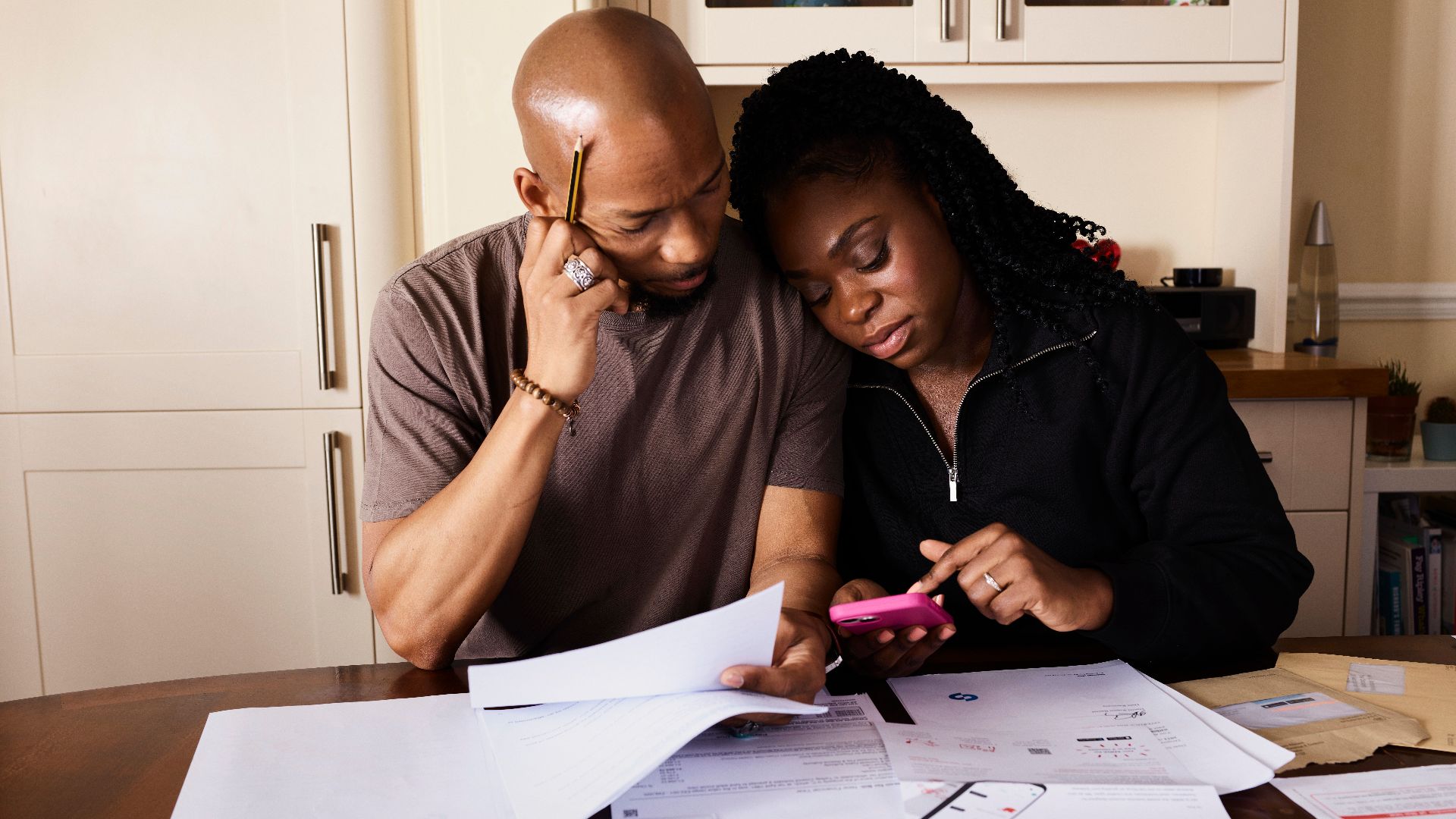 A couple sits at a table managing domestic finances, evaluating documents and using a smartphone.