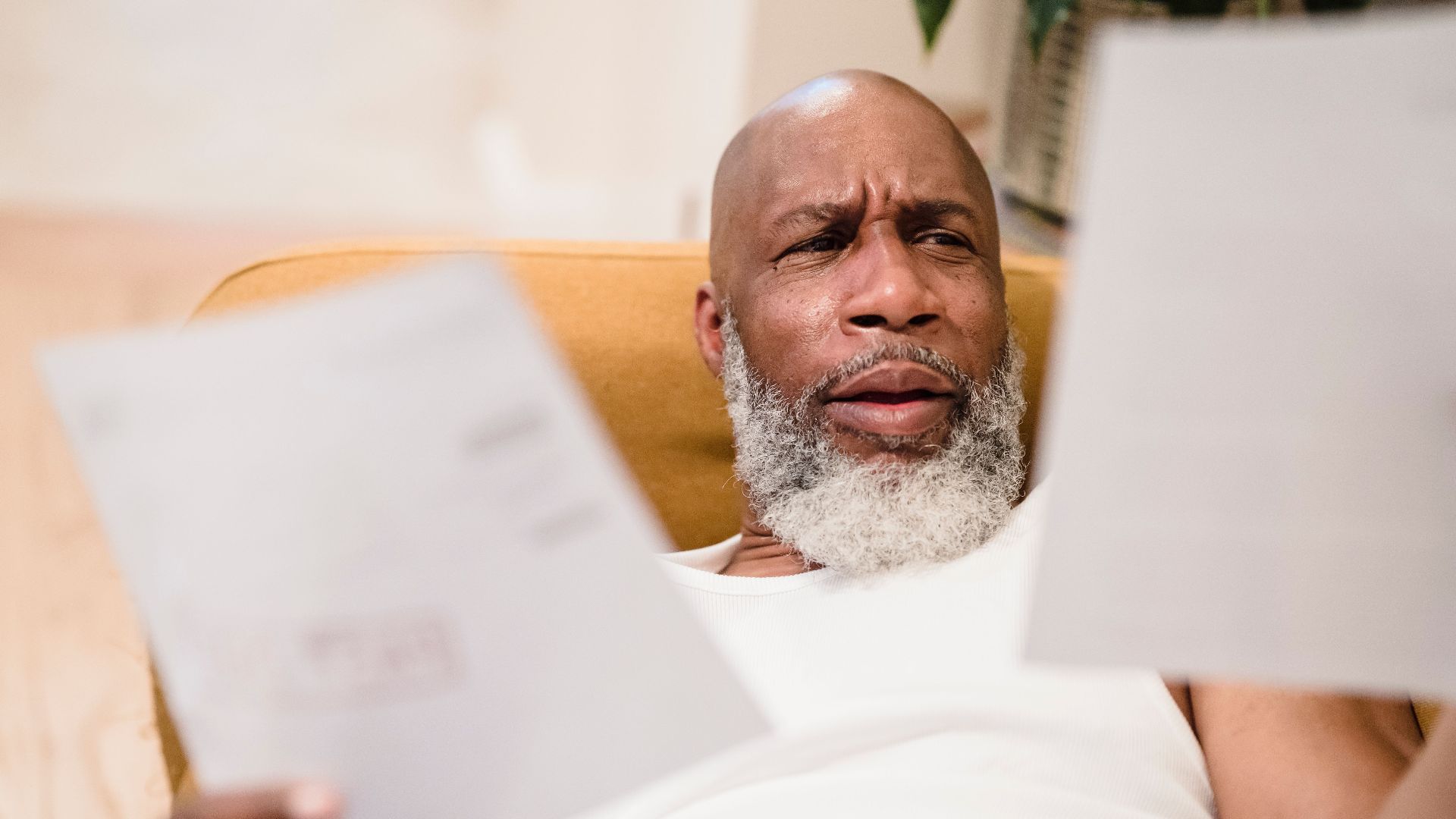 A middle-aged man looks concerned as he reviews household bills, sitting in a comfortable chair indoors.