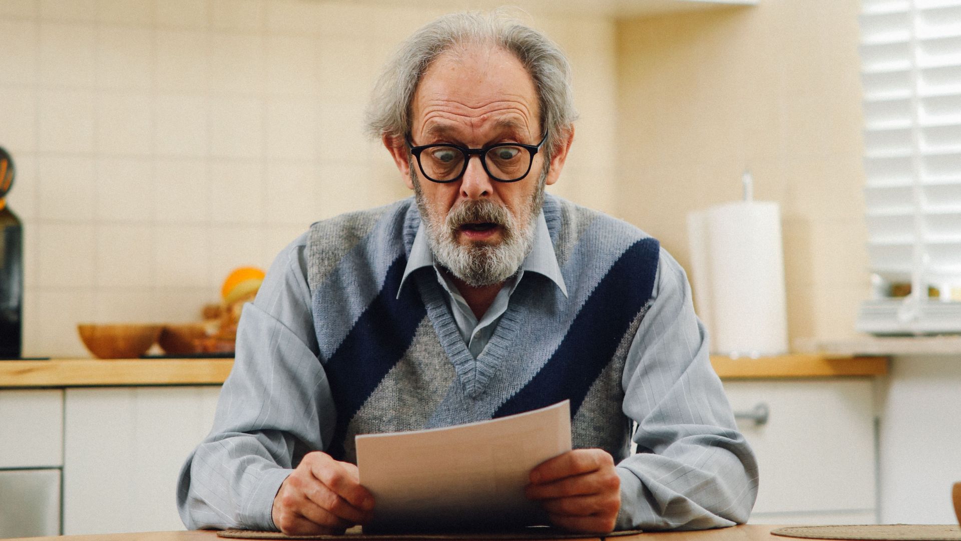 Senior man with eyeglasses reading a document at kitchen table, reflecting surprise.