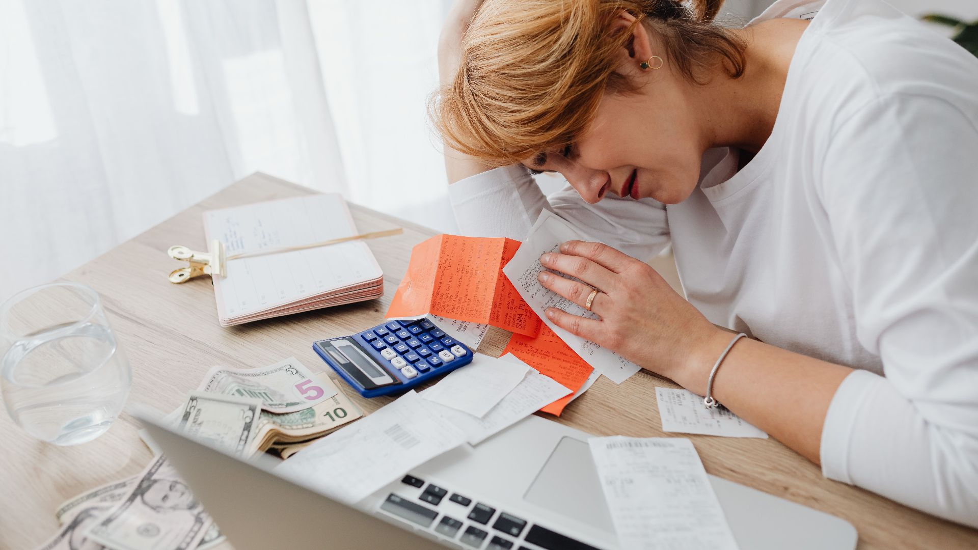 A woman overwhelmed with finances, surrounded by bills, calculator, and cash at home.