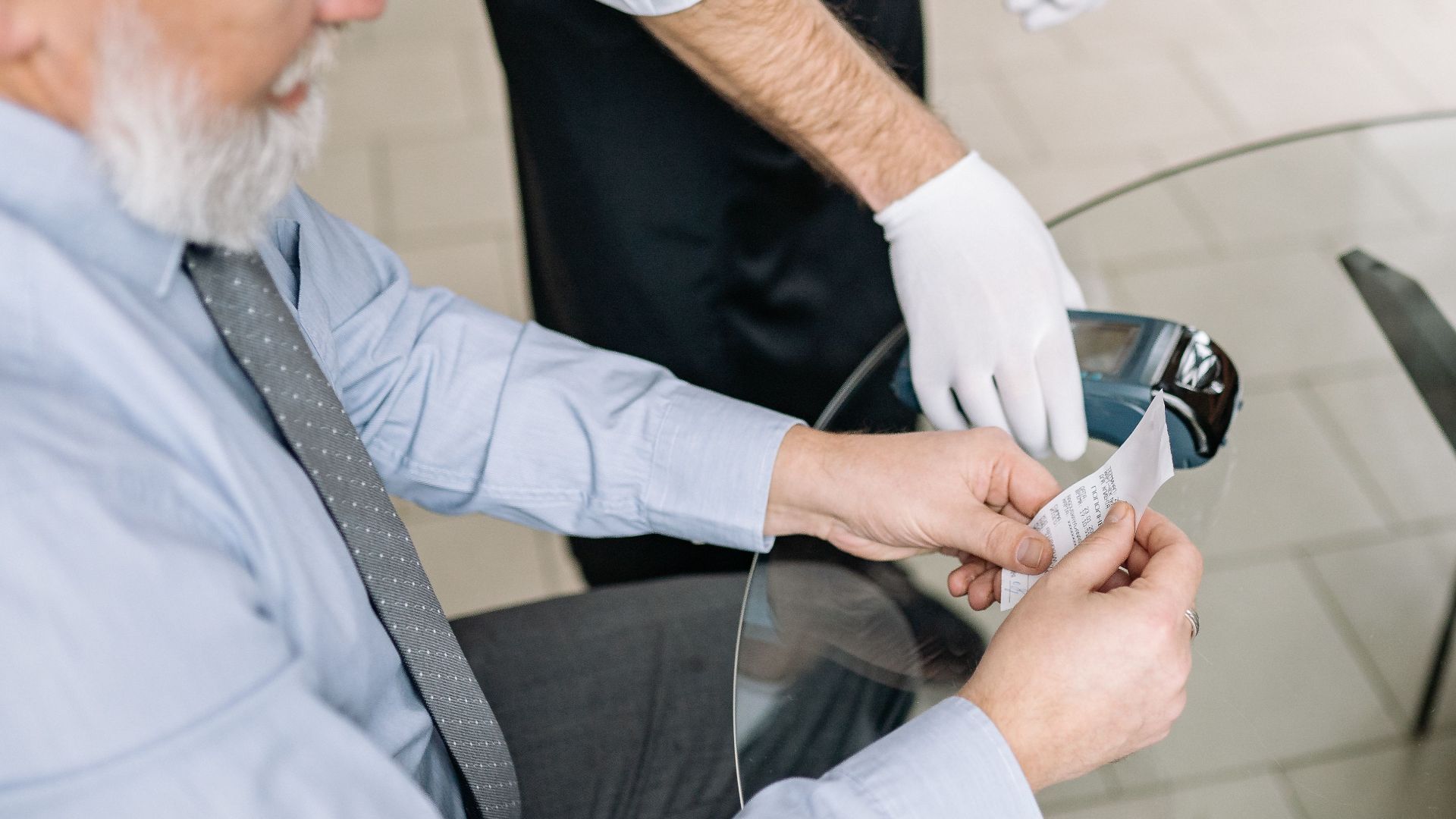 Senior man making a payment via credit card with a waiter in gloves assisting.