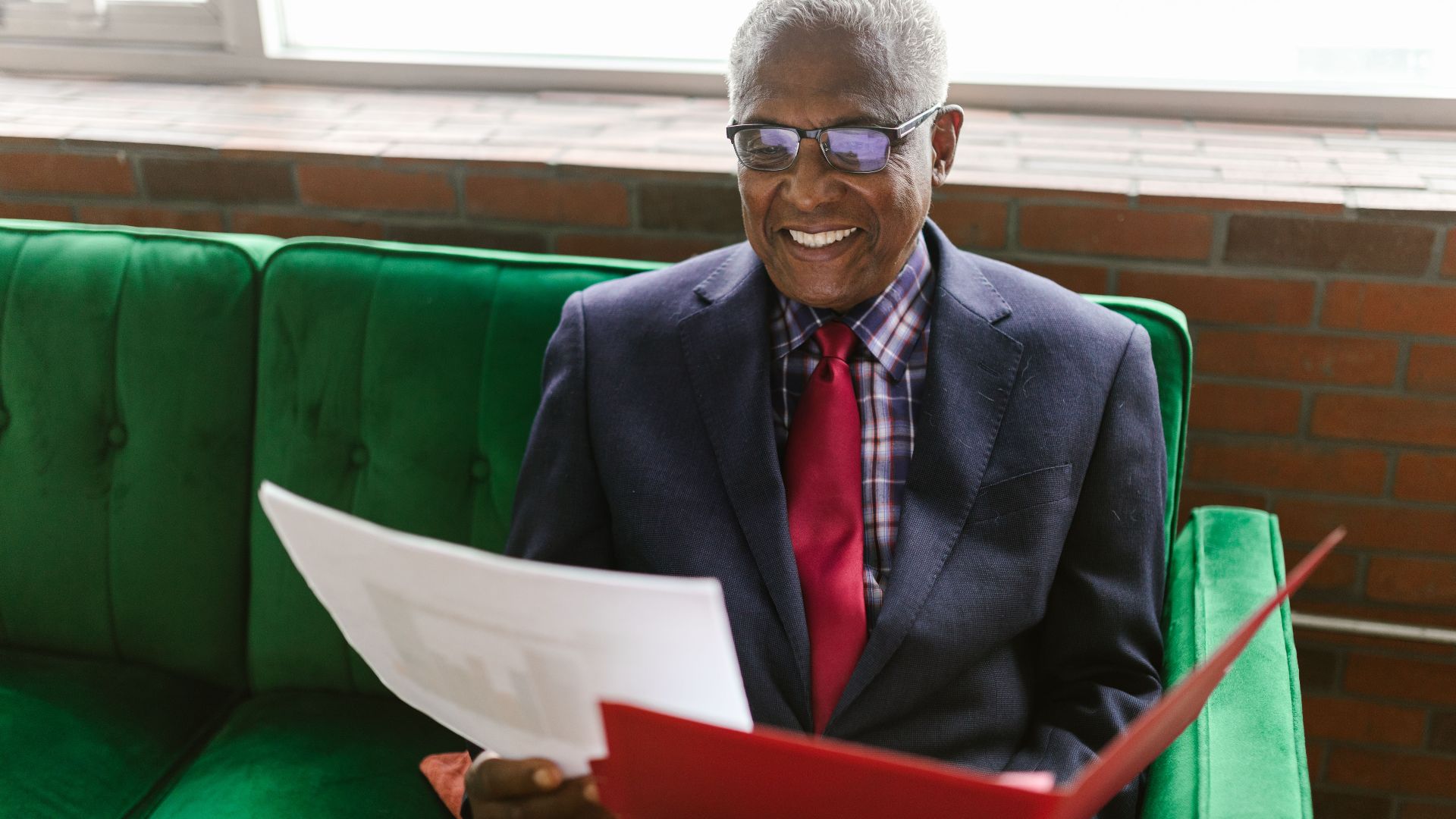 Elderly man in a blue suit sits on a green sofa, reviewing paperwork with a smile.