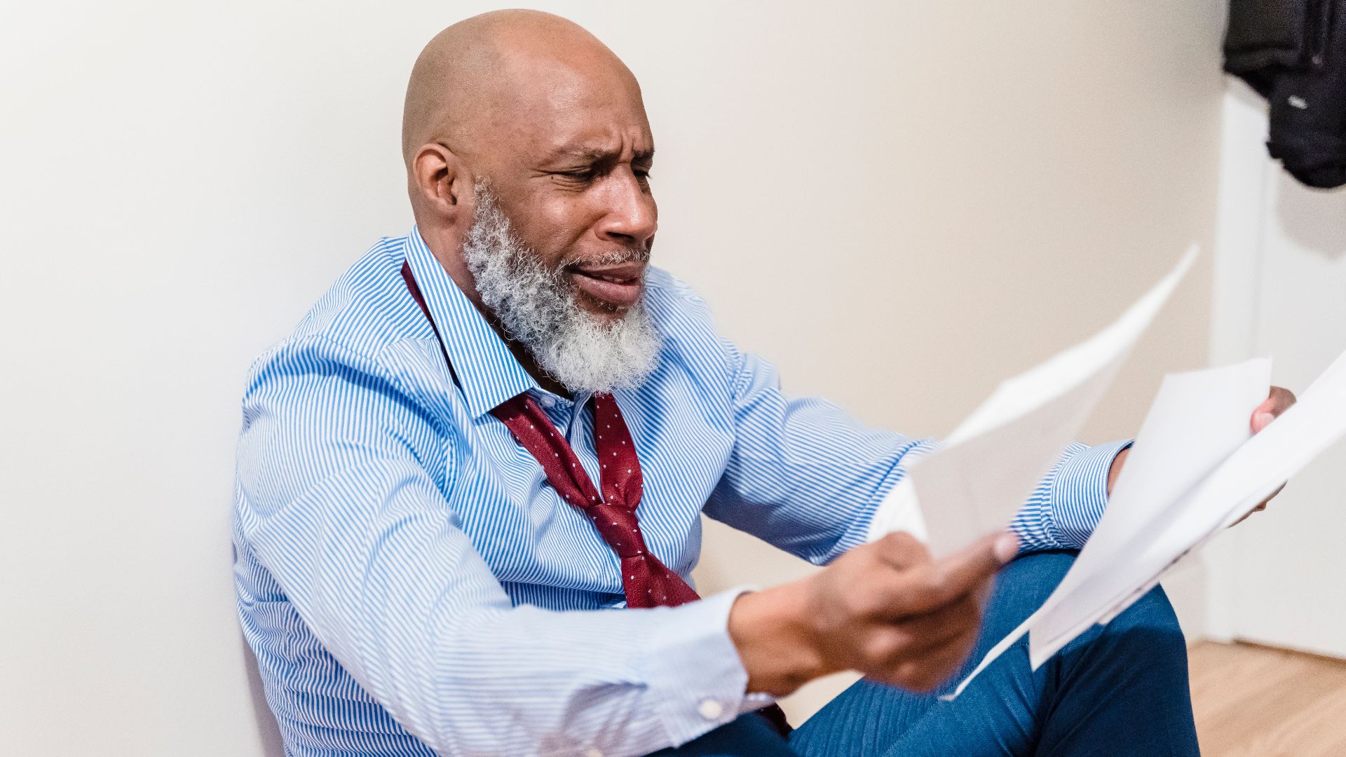 An adult man with a beard looks distressed while sitting on the floor, examining papers.