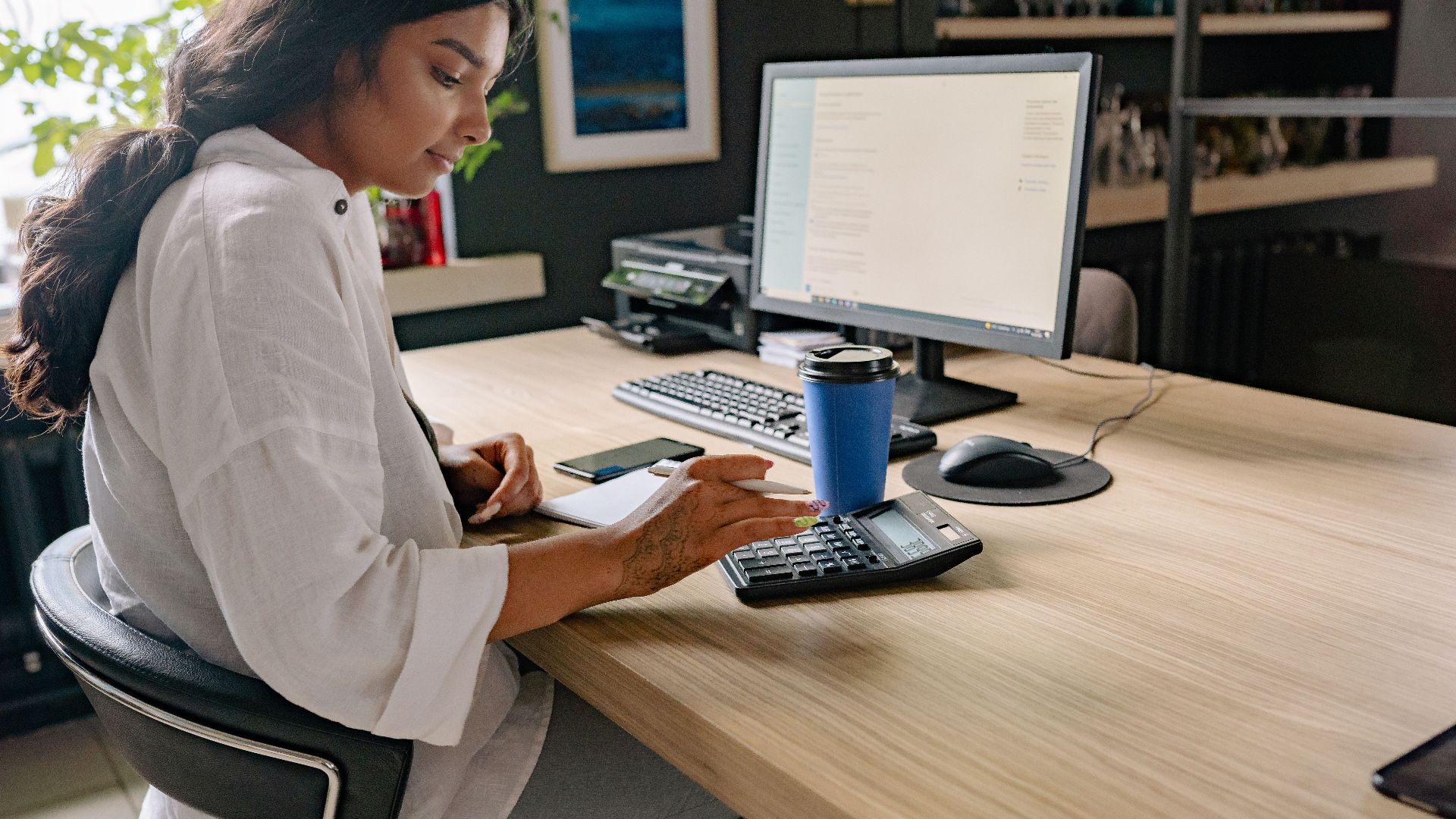 A professional woman using a calculator and computer at an office desk, focused on her work.