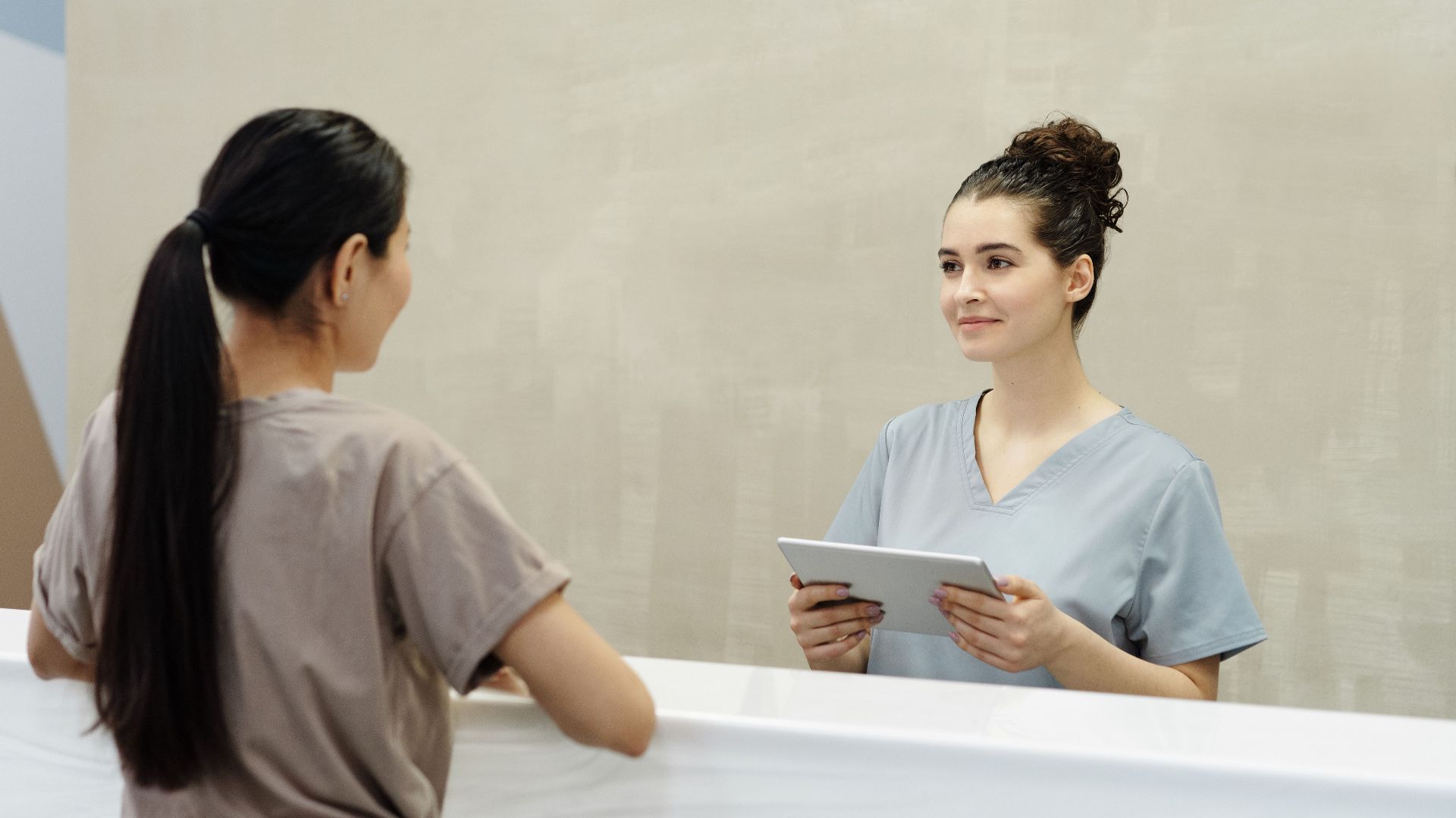 A female receptionist using a tablet to assist a patient at a modern indoor reception desk.