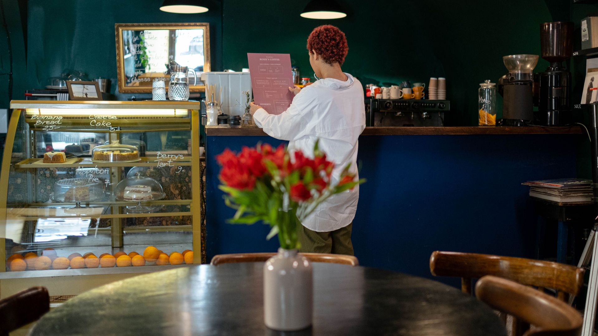 A barista reads an order in a cozy café with a pastry display and warm lighting.