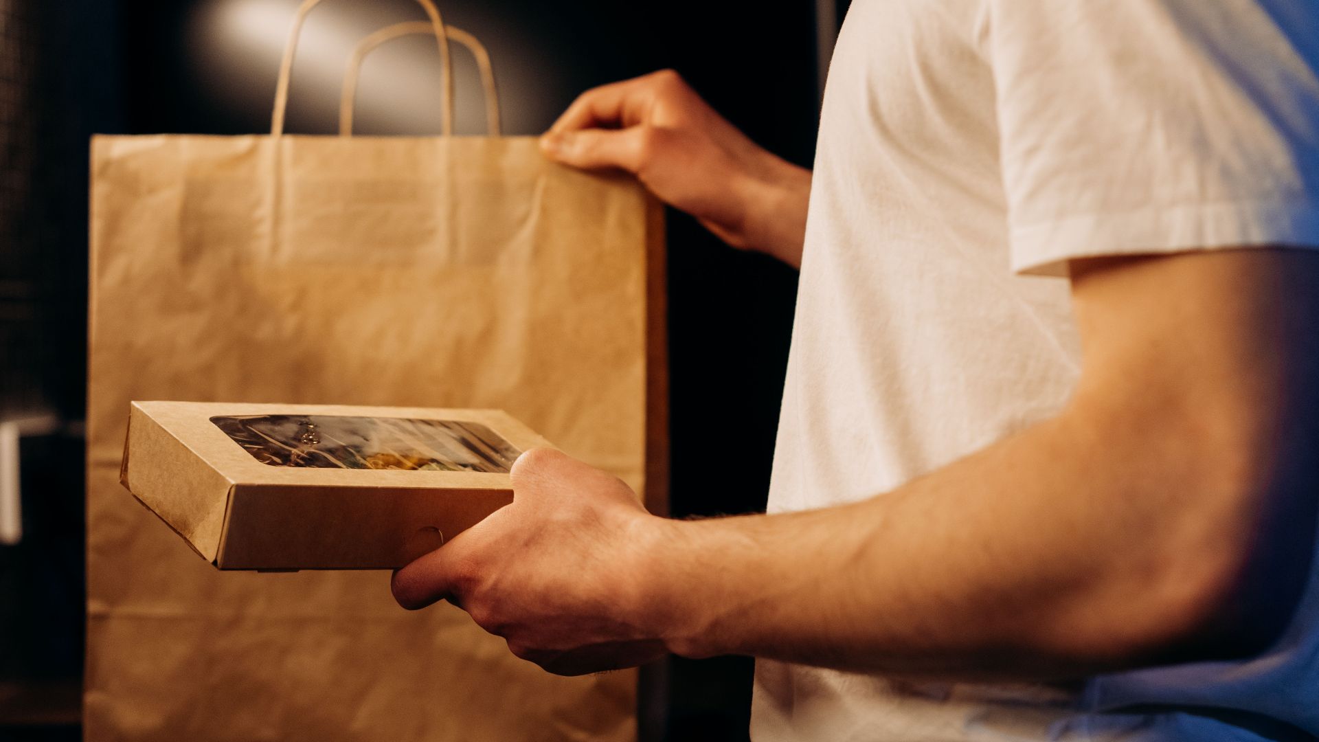 A person presenting a food delivery package and paper bag at home.