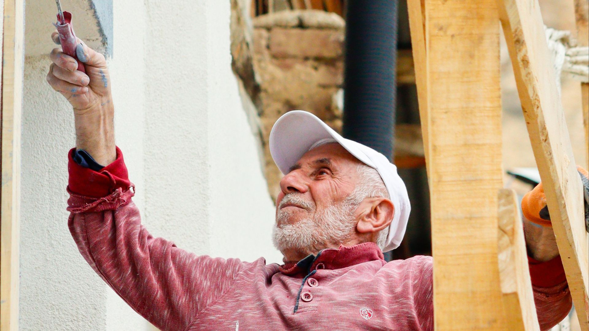 Elderly man wearing a cap paints an exterior wall while seated on scaffolding.