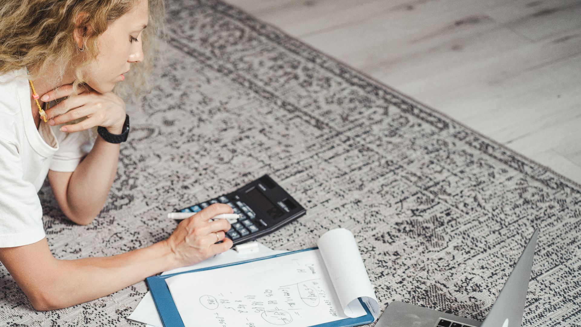 A woman calculates finances at home using a laptop, calculator, and documents on a rug.