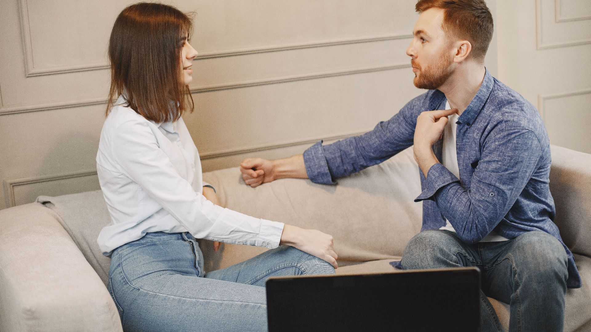 A young couple engaged in a conversation while sitting on a beige couch indoors.