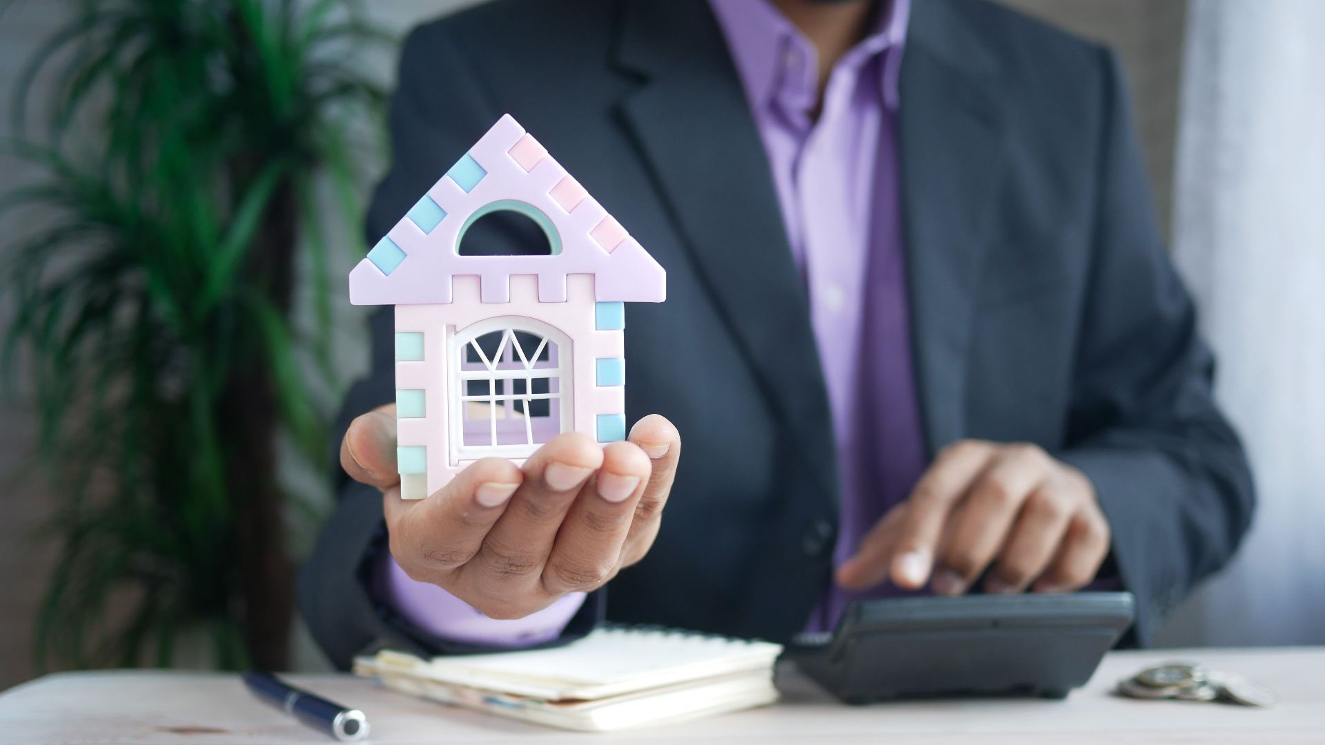 Businessman in suit holds model house, calculates real estate investments.