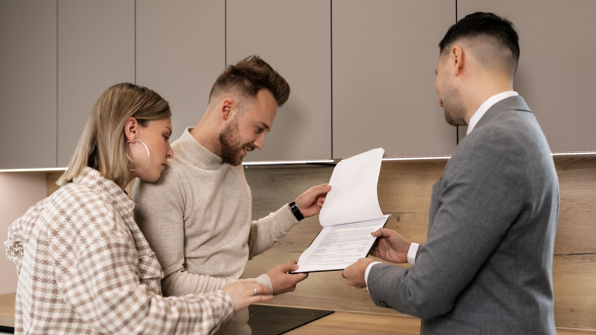 A couple reviews real estate documents with an agent in a modern indoor setting, discussing a potential property purchase.