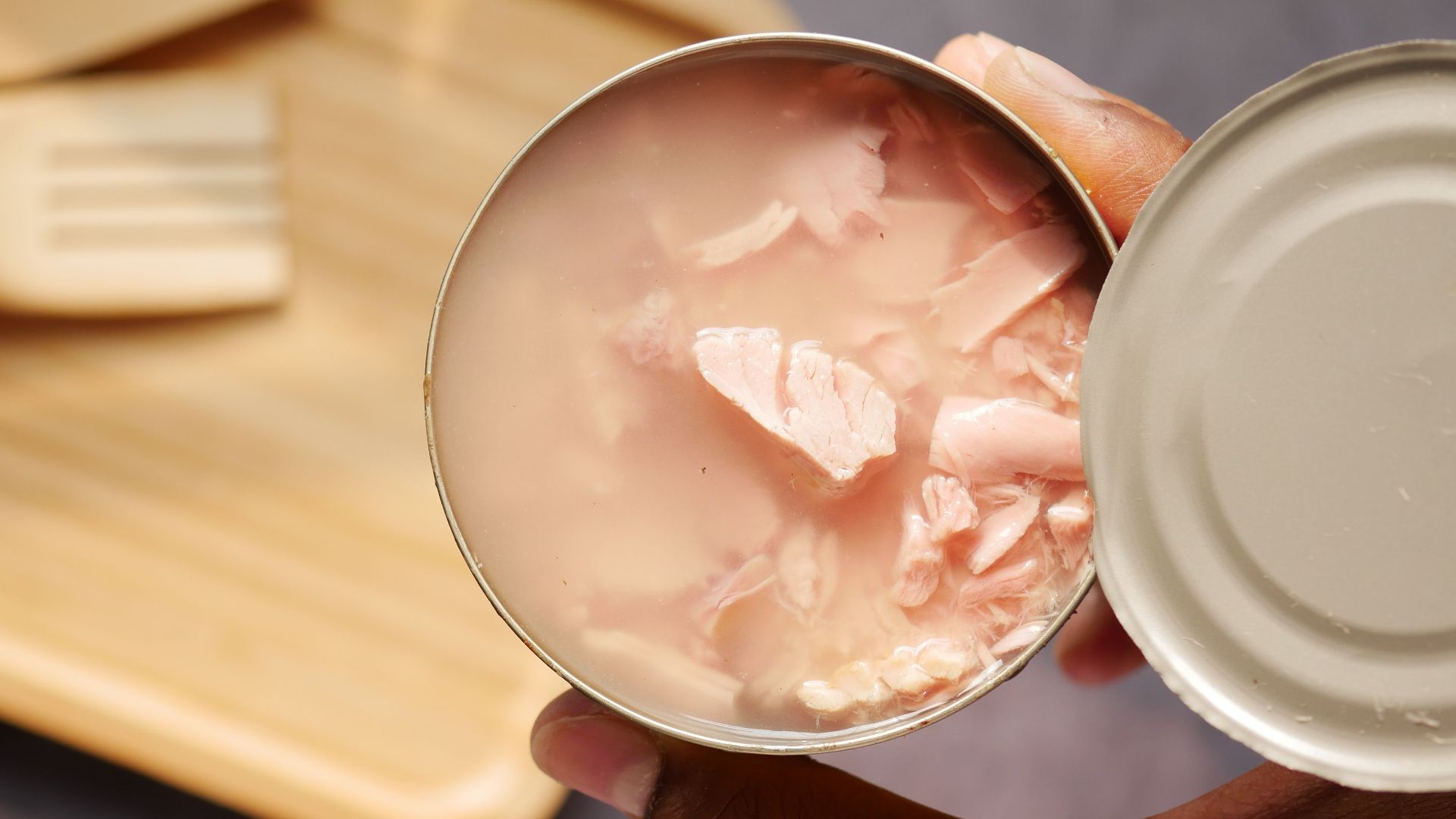 A close-up of an opened canned tuna held over a wooden cutting board and utensils.