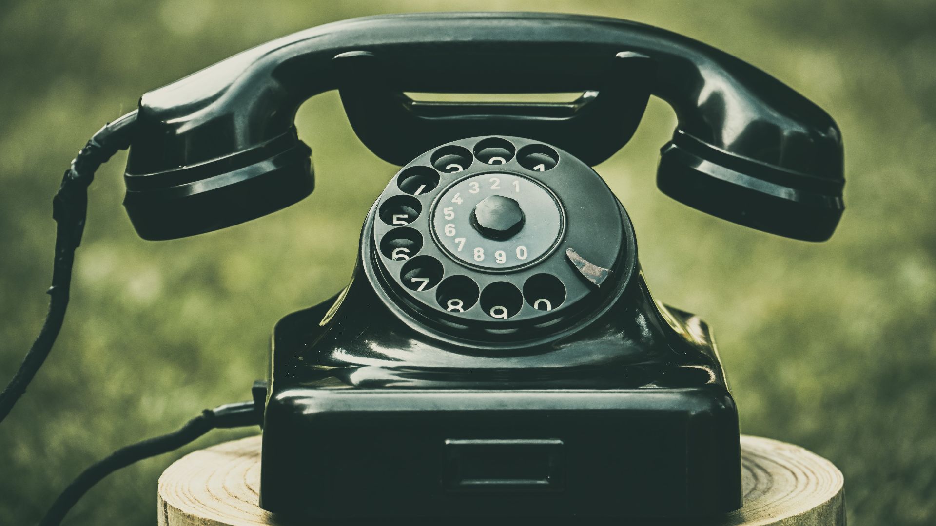 A classic black rotary dial telephone placed outdoors on a wooden stump, evoking nostalgia.