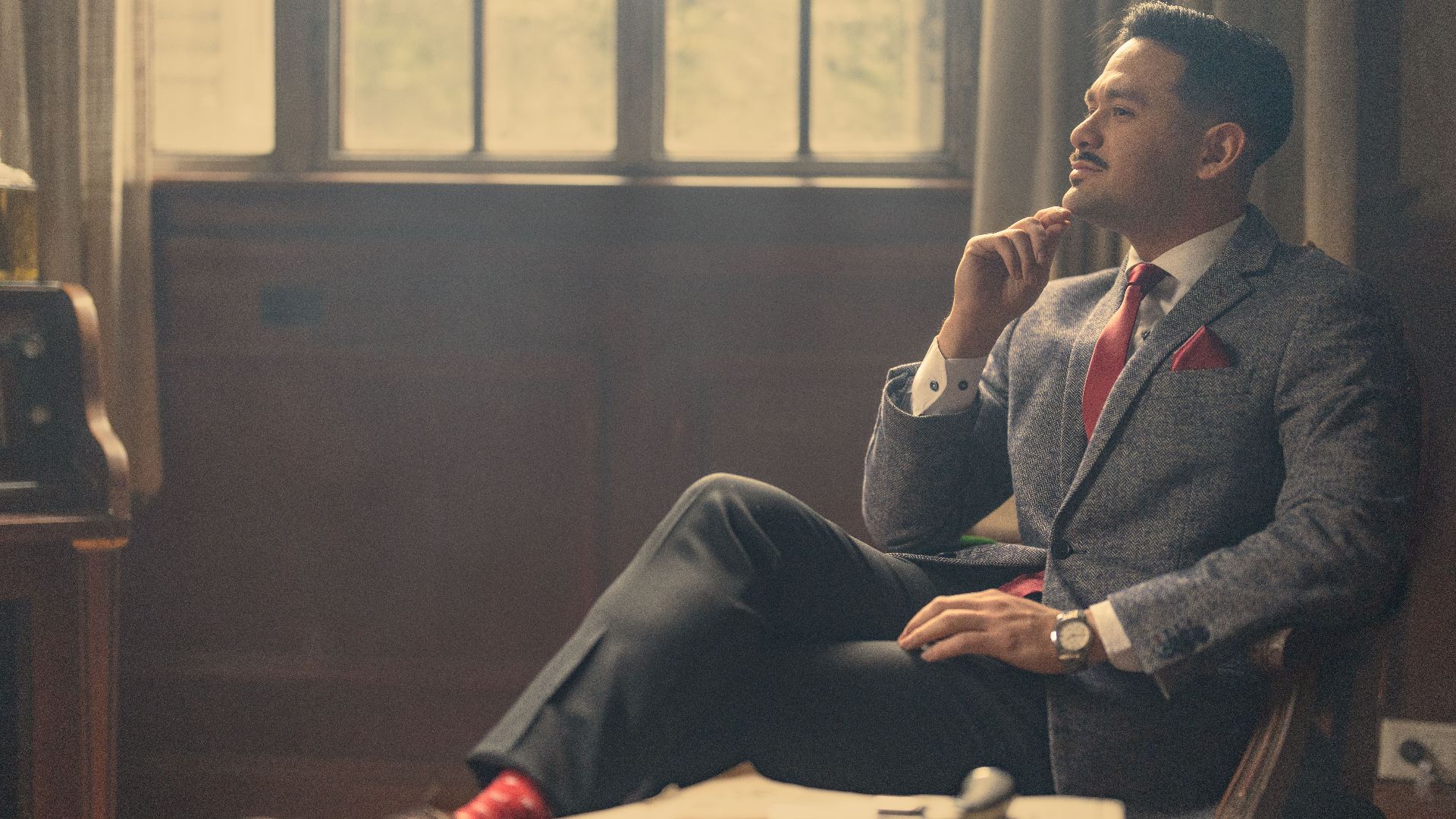 A stylish man in a suit sits thoughtfully in a vintage office, exuding confidence and elegance.