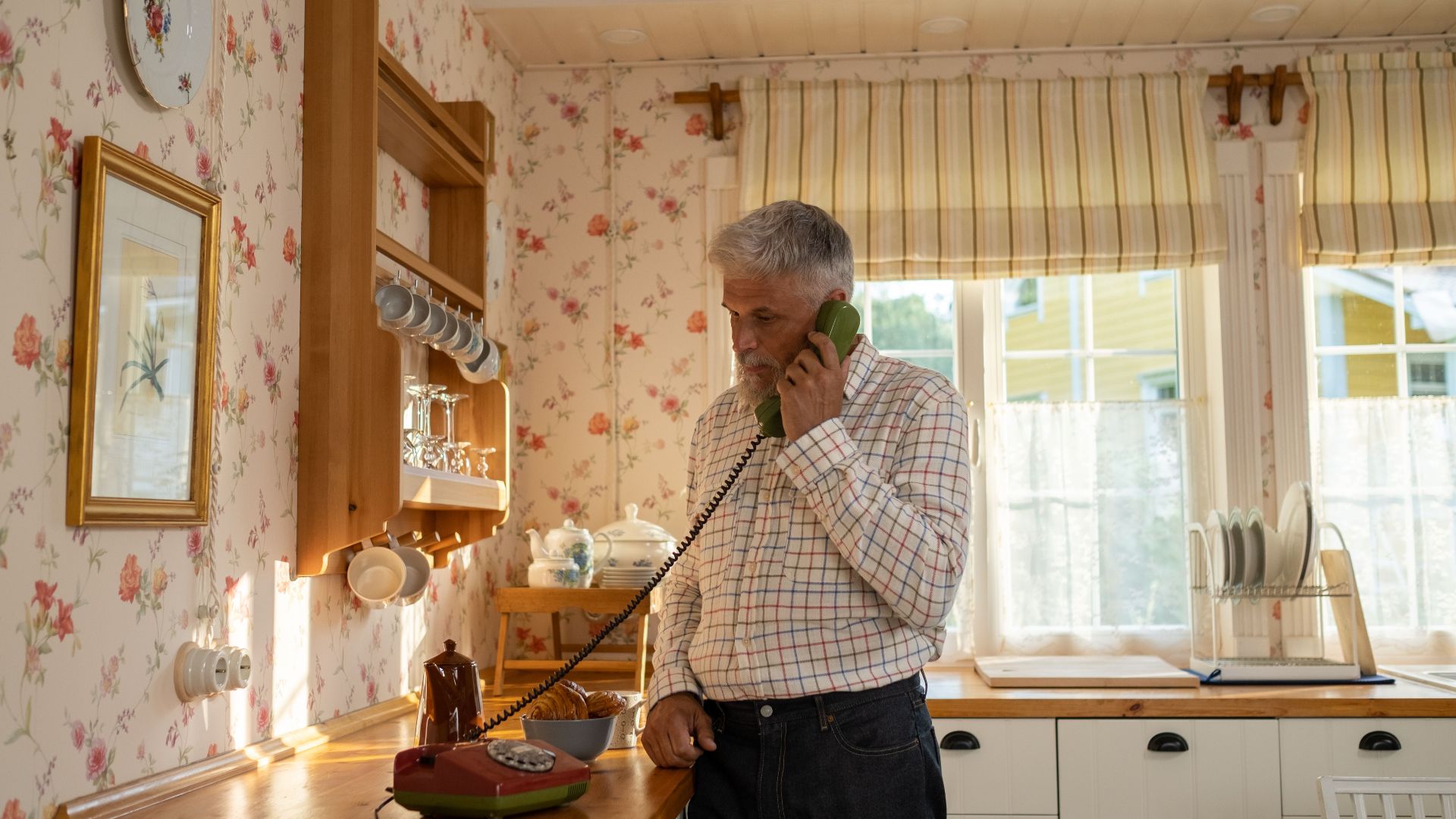 Senior man talks on a vintage rotary phone in a retro-styled kitchen with floral wallpaper and classic decor.