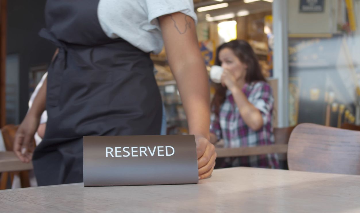 Close up of waiter putting reserved plate on table in restaurant with happy couple drinking coffee on date on blurred background. Restaurant manager placing reserved table sign