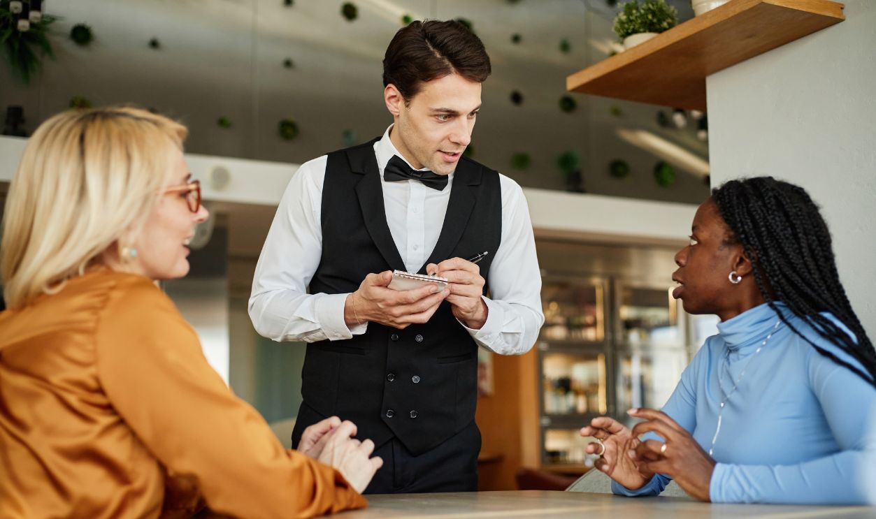 Caucasian young adult waiter taking order from diverse women sitting at restaurant table, both women engaging in conversation with waiter