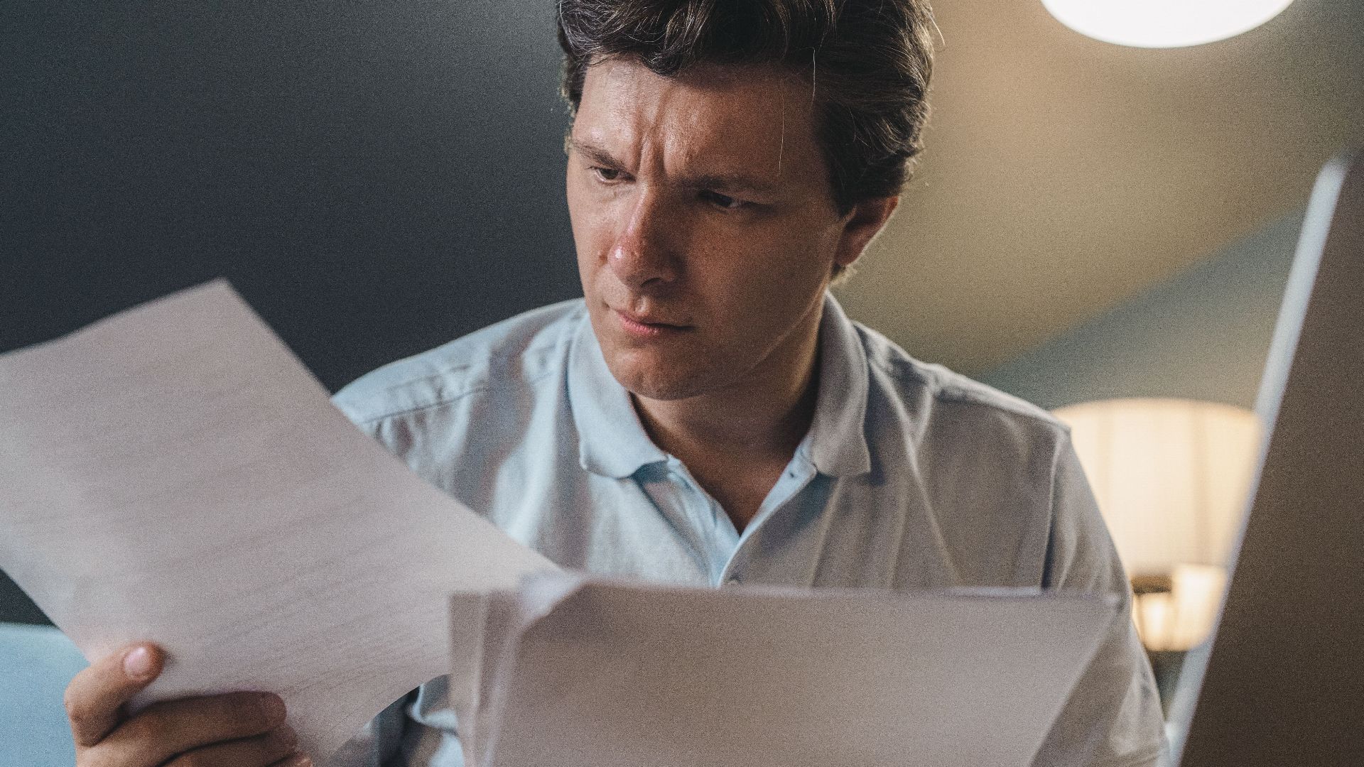 A man intently examines papers, seated indoors under warm lighting, focusing on his work.
