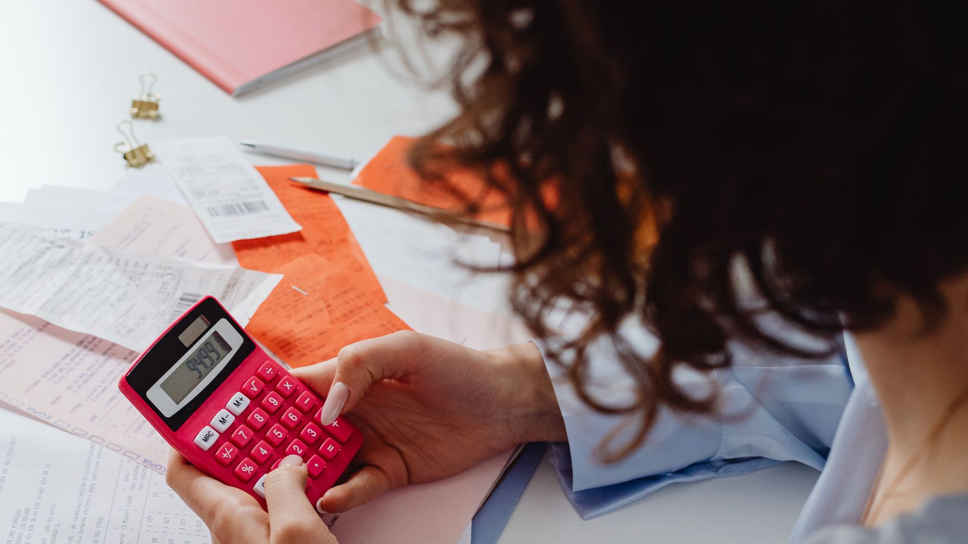 Adult woman working with receipts and a bright pink calculator at a desk, focusing on finances.