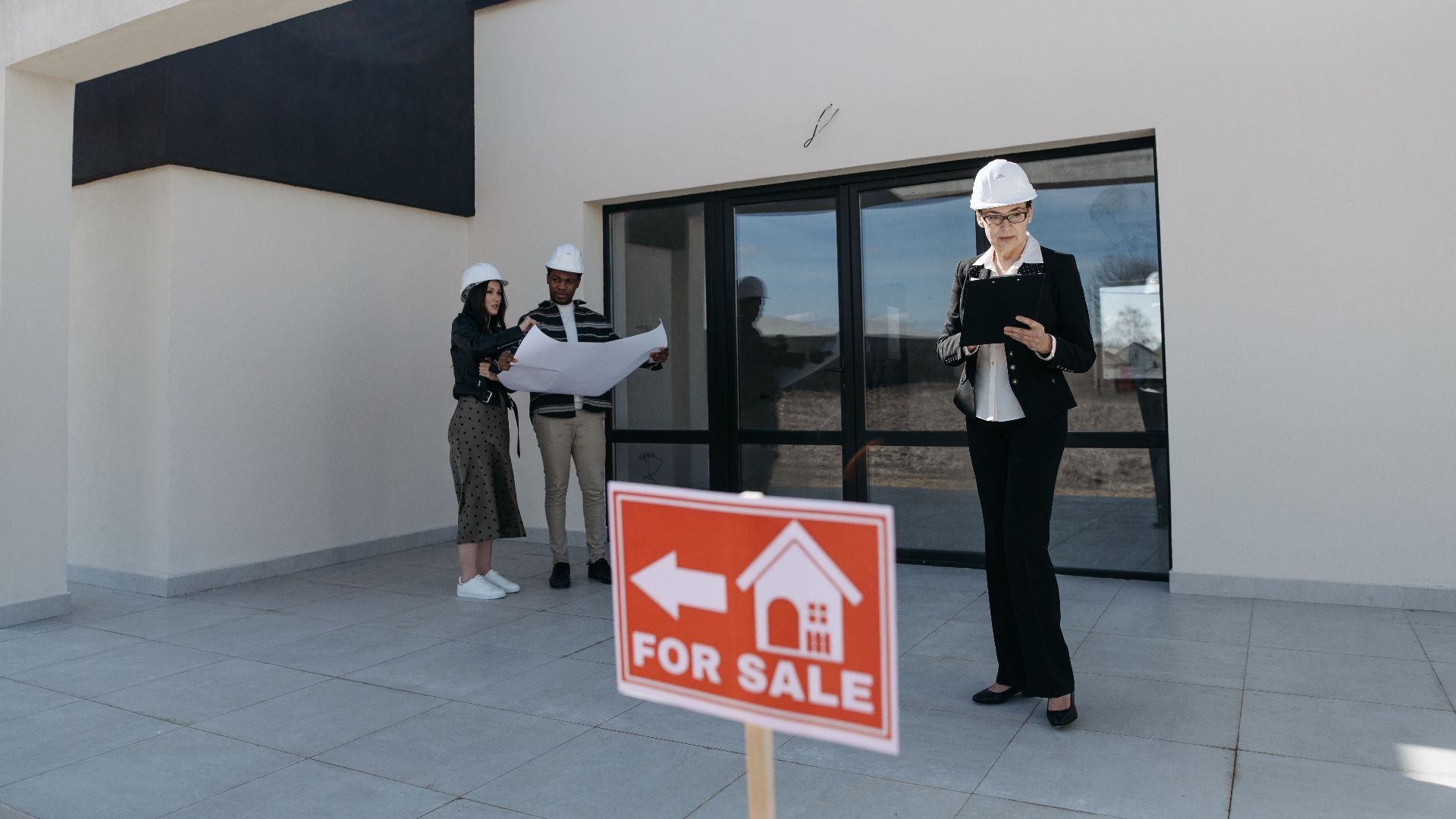 Real estate agents in hard hats reviewing plans beside a house for sale.