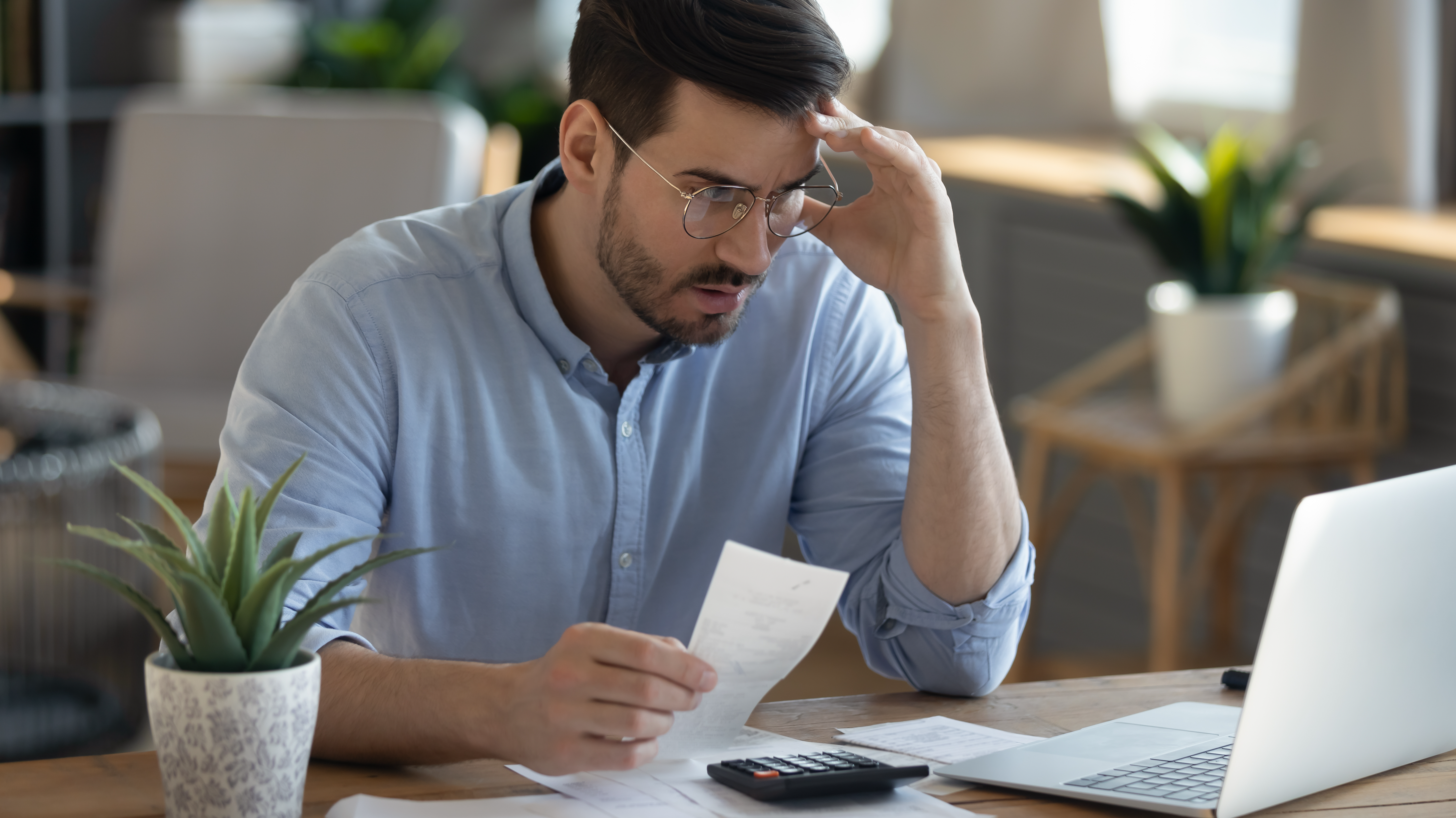 Man sitting at desk looks at laptop screen holding receipt feels desperate after calculating expenses.