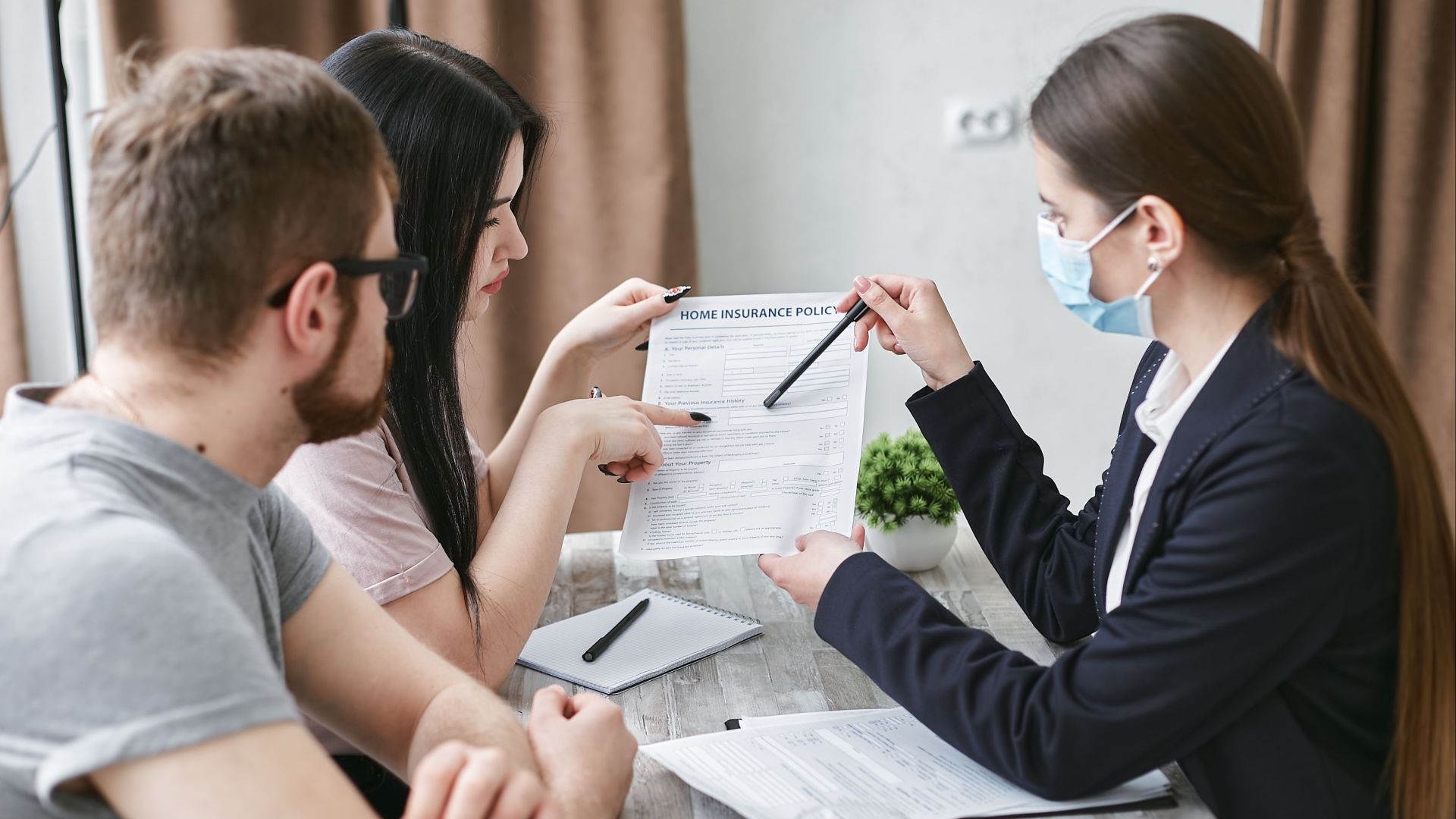 Three adults discuss a home insurance policy at a meeting table indoors.