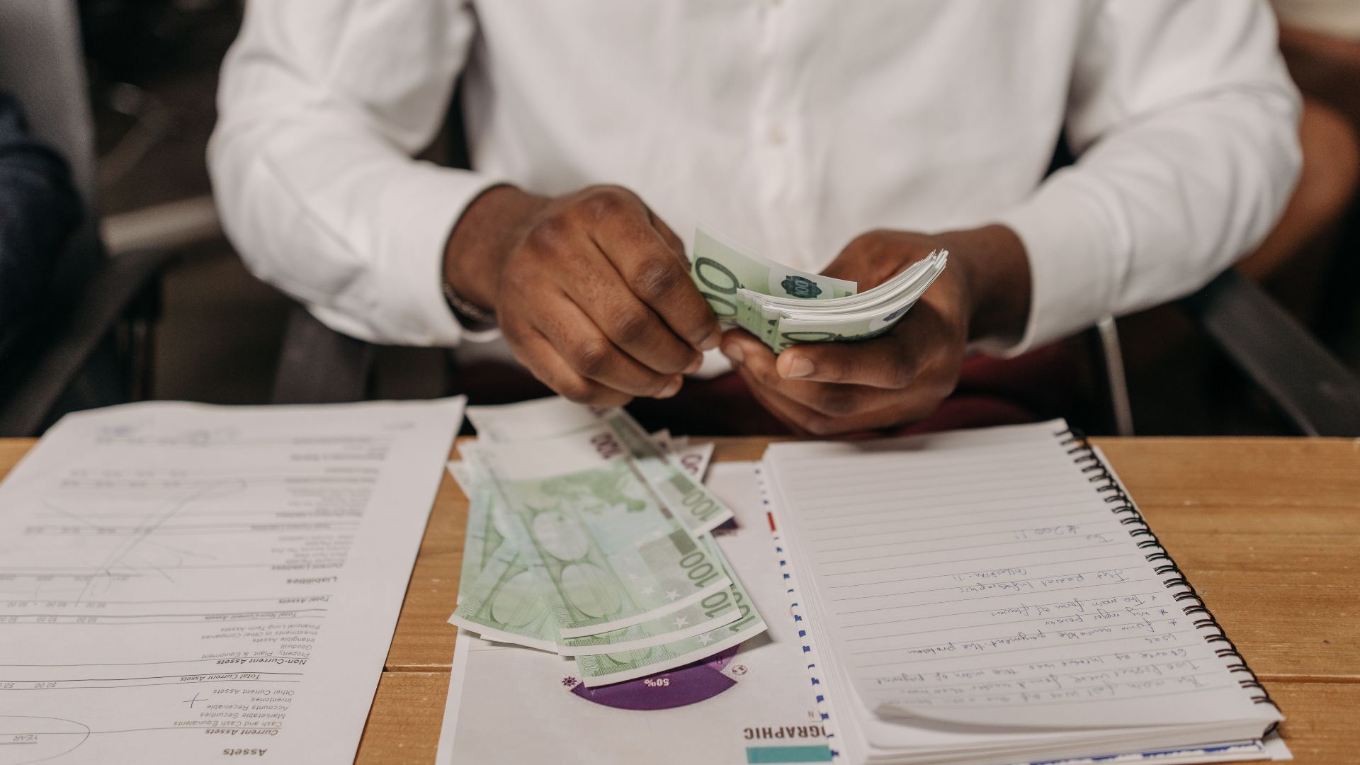 African businessman counting euro bills at a desk with documents and a notebook, highlighting finance theme.