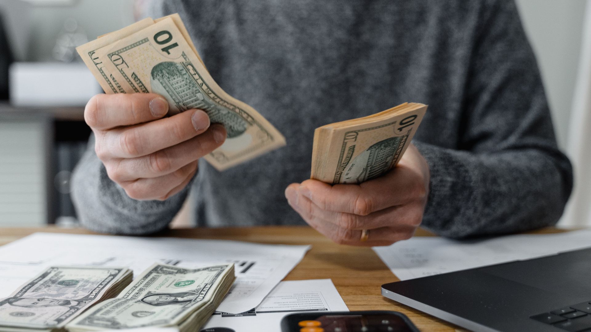Close-up of person counting cash with a calculator and paperwork on a desk.