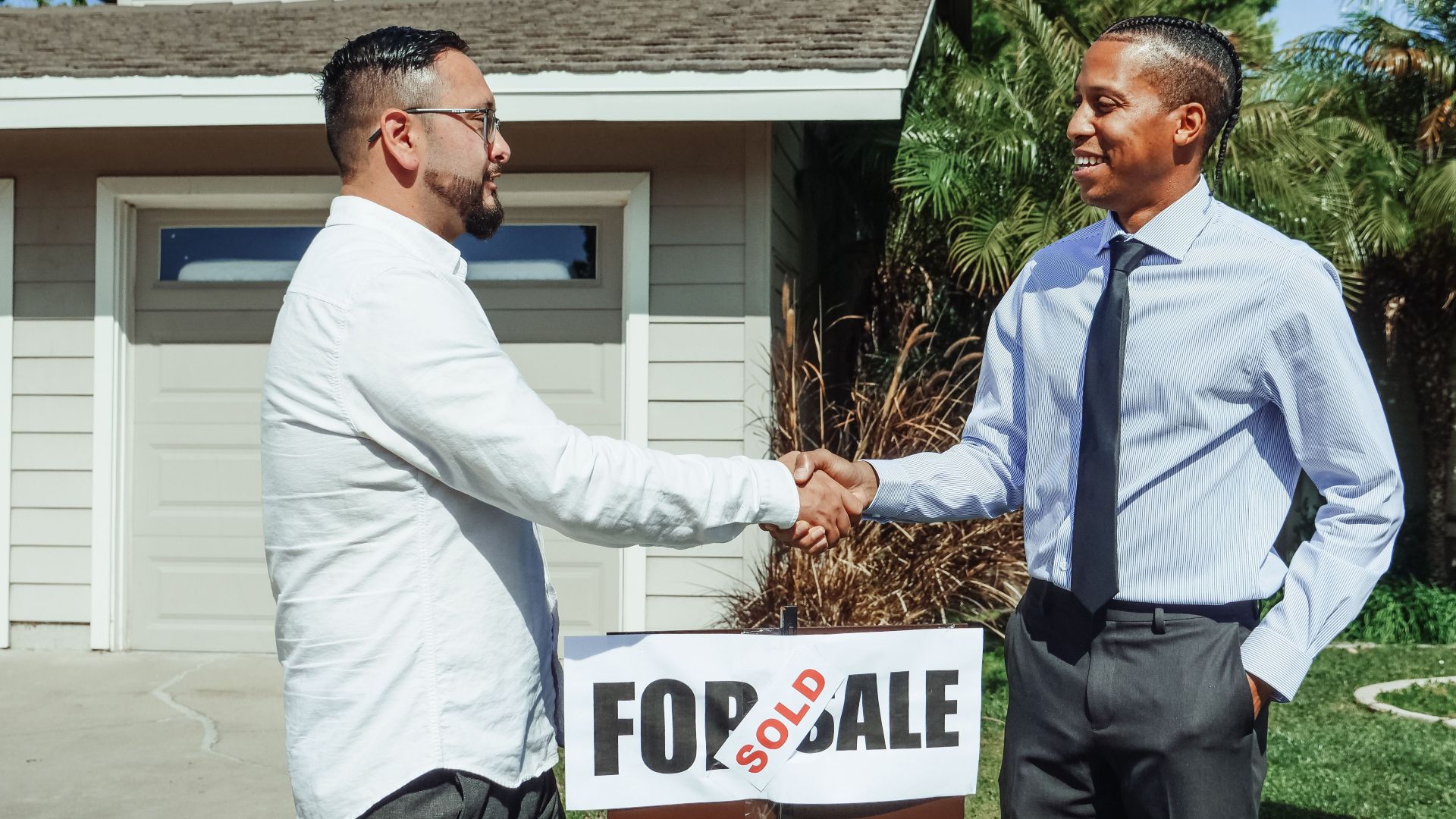 Two men shaking hands in front of house sold sign, sealing real estate deal outdoors.