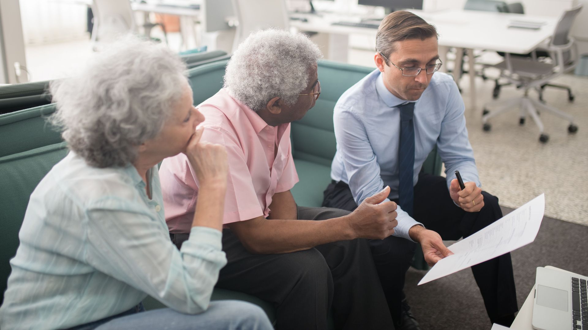 Business professional consults elderly clients in an office setting. Collaborative discussion, paperwork visible.