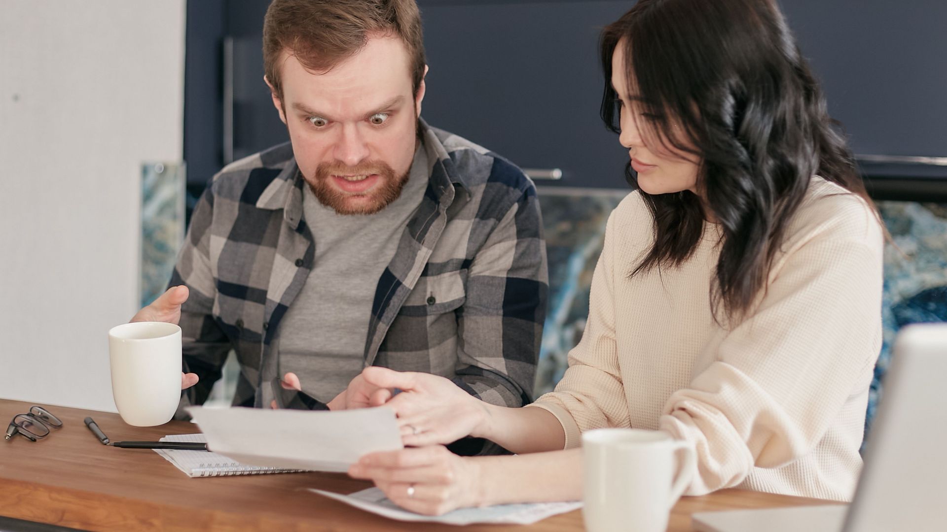 A couple shows astonishment as they review documents together at a wooden desk indoors.