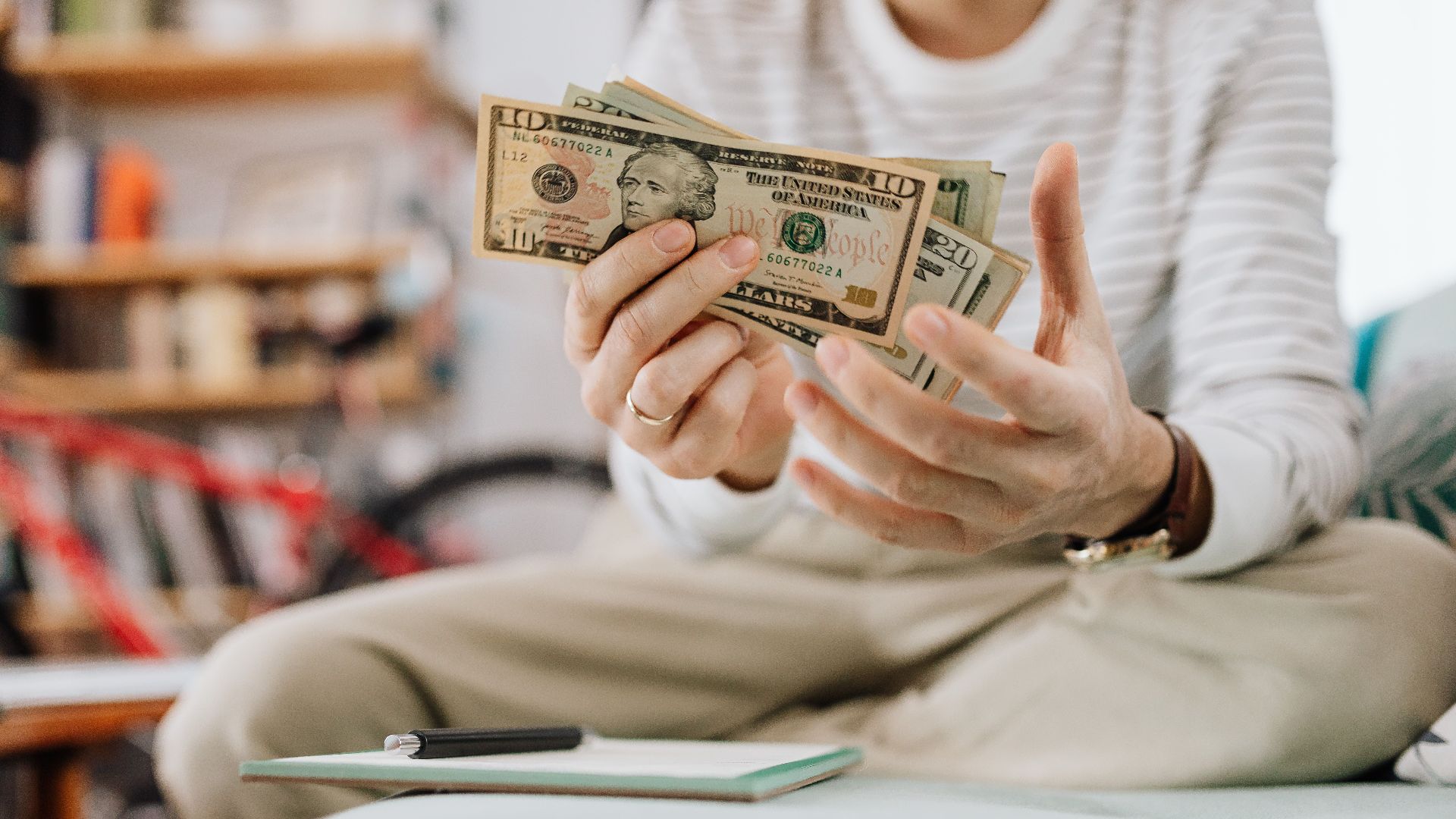 Man wearing casual clothes counts dollar bills while sitting on a sofa indoors.