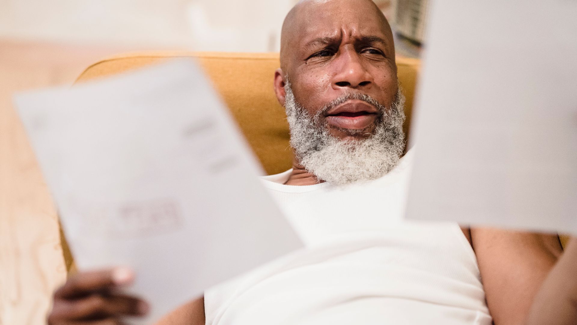 A middle-aged man looks concerned as he reviews household bills, sitting in a comfortable chair indoors.