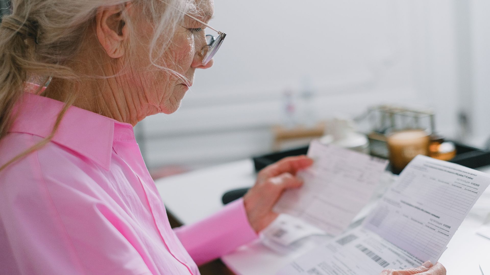 Elderly woman in a pink shirt reviewing paperwork at home.