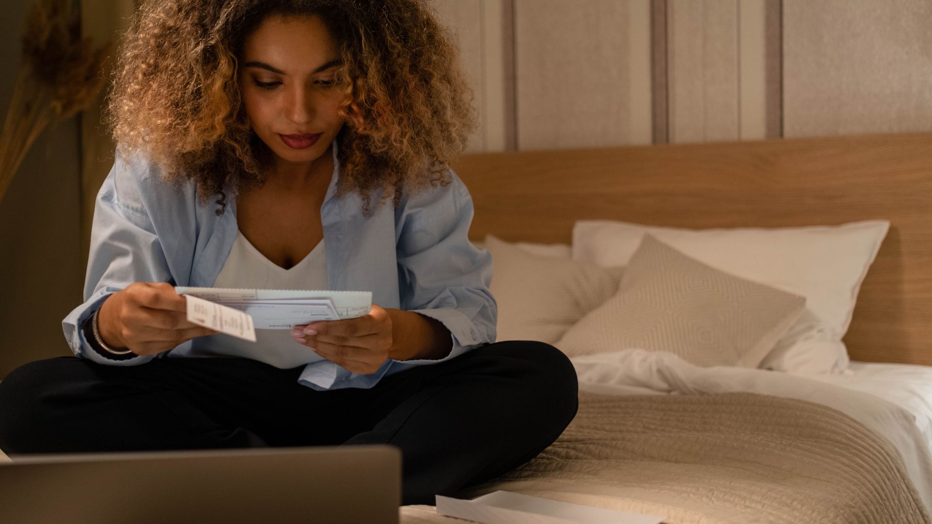 Woman sitting on bed reading documents with laptop, highlighting home office setting.