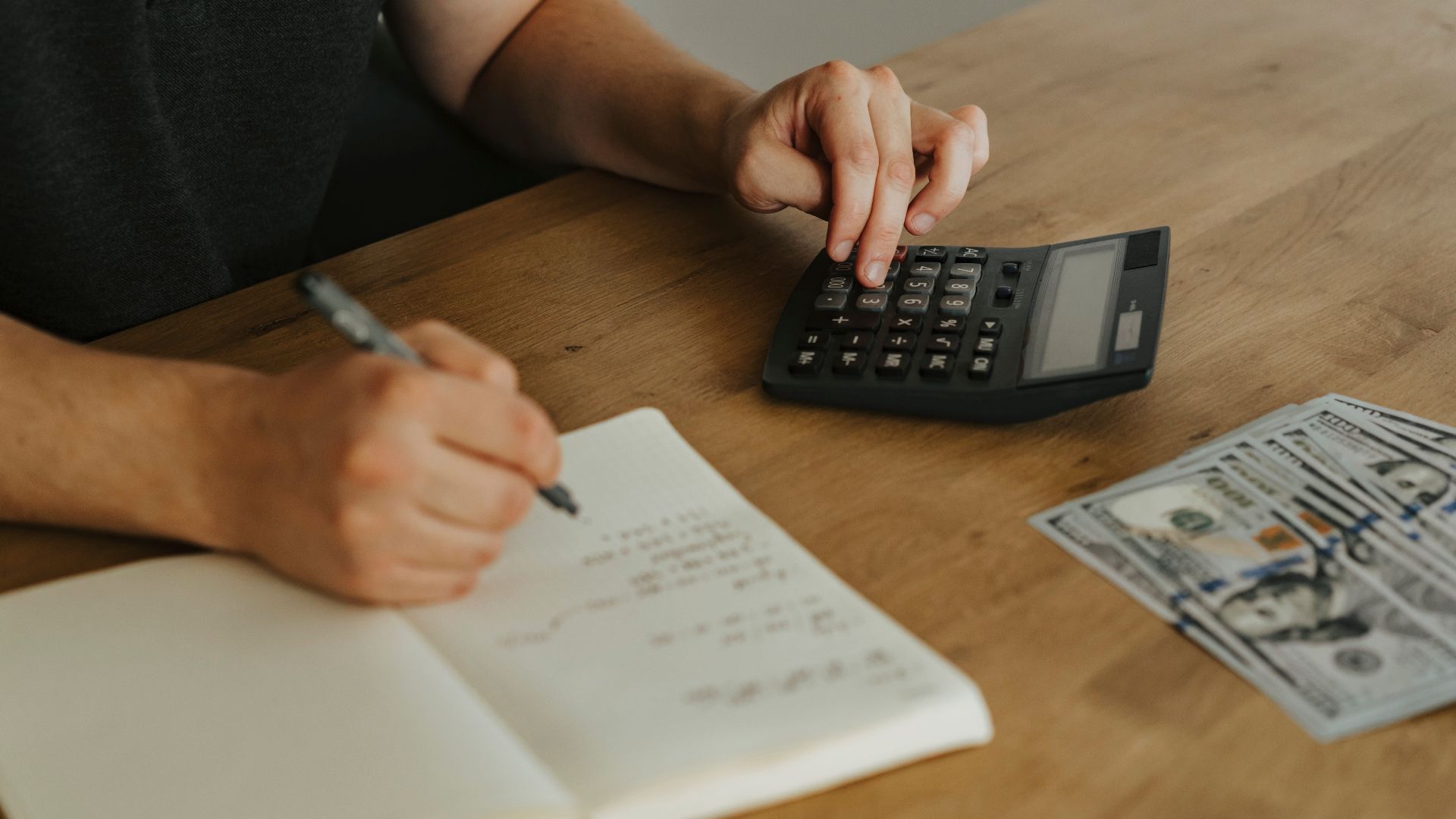 Close-up of hands using a calculator with notebook and cash on a wooden table.