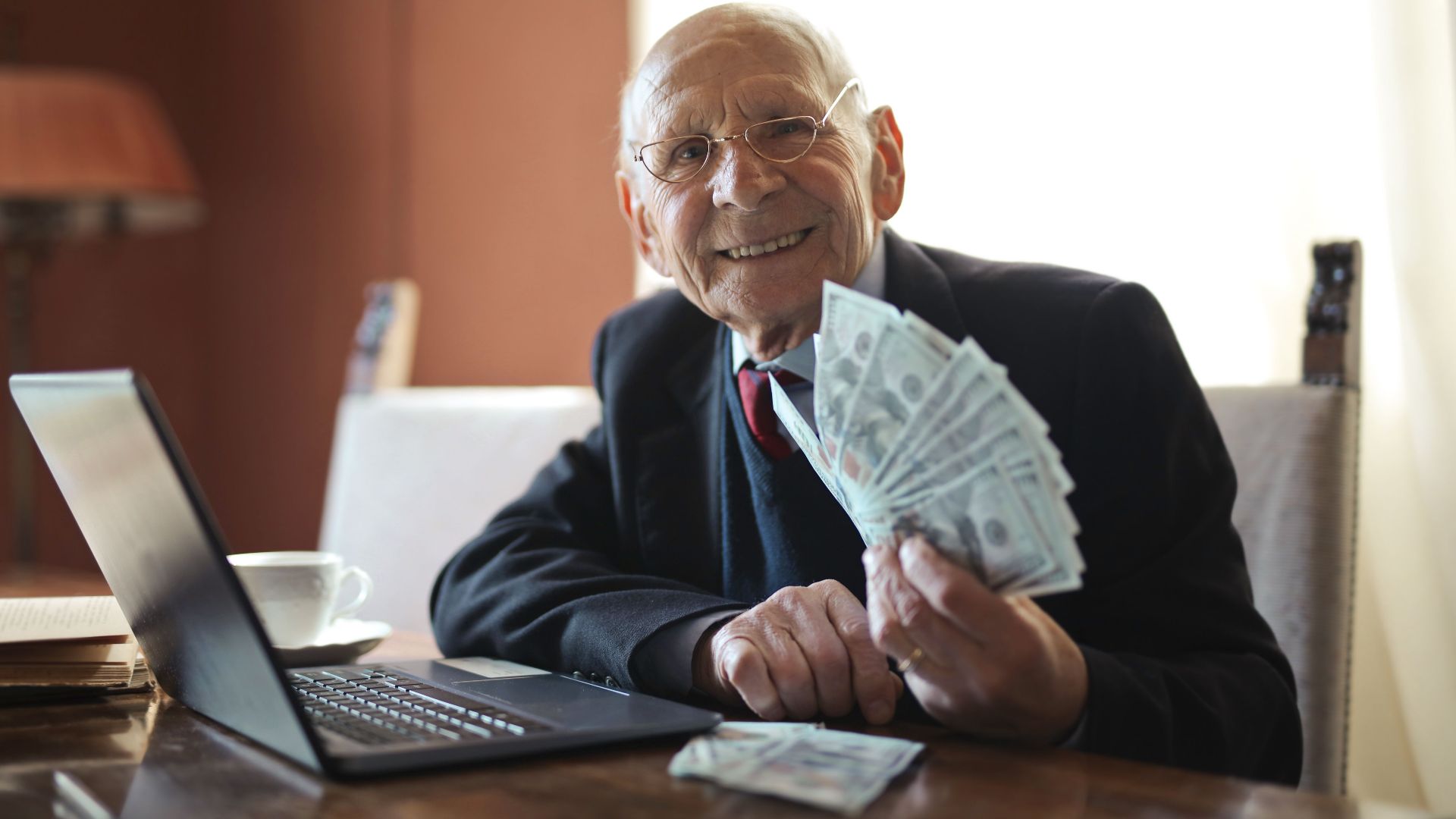 Positive senior businessman in formal suit and eyeglasses holding bunch of bills while working on laptop at table and looking at camera