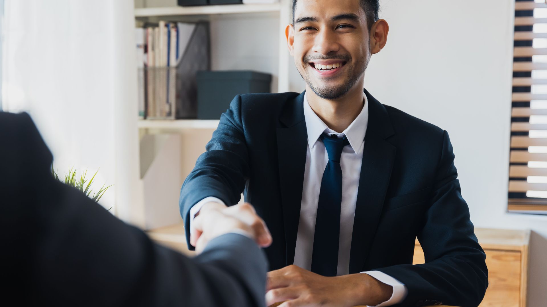Confident businessman in suit shaking hands at office desk, symbolizing successful partnership.