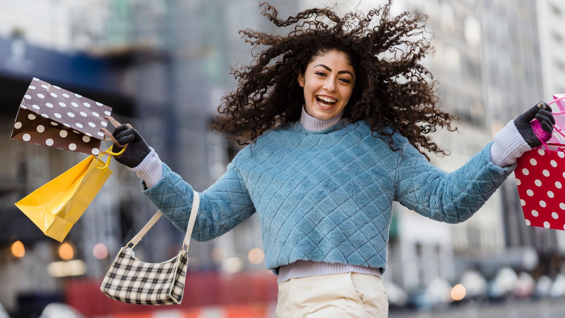 Cheerful female shopper with curly hair holding colorful paper bags in raised hands and happily laughing and looking at camera while jumping