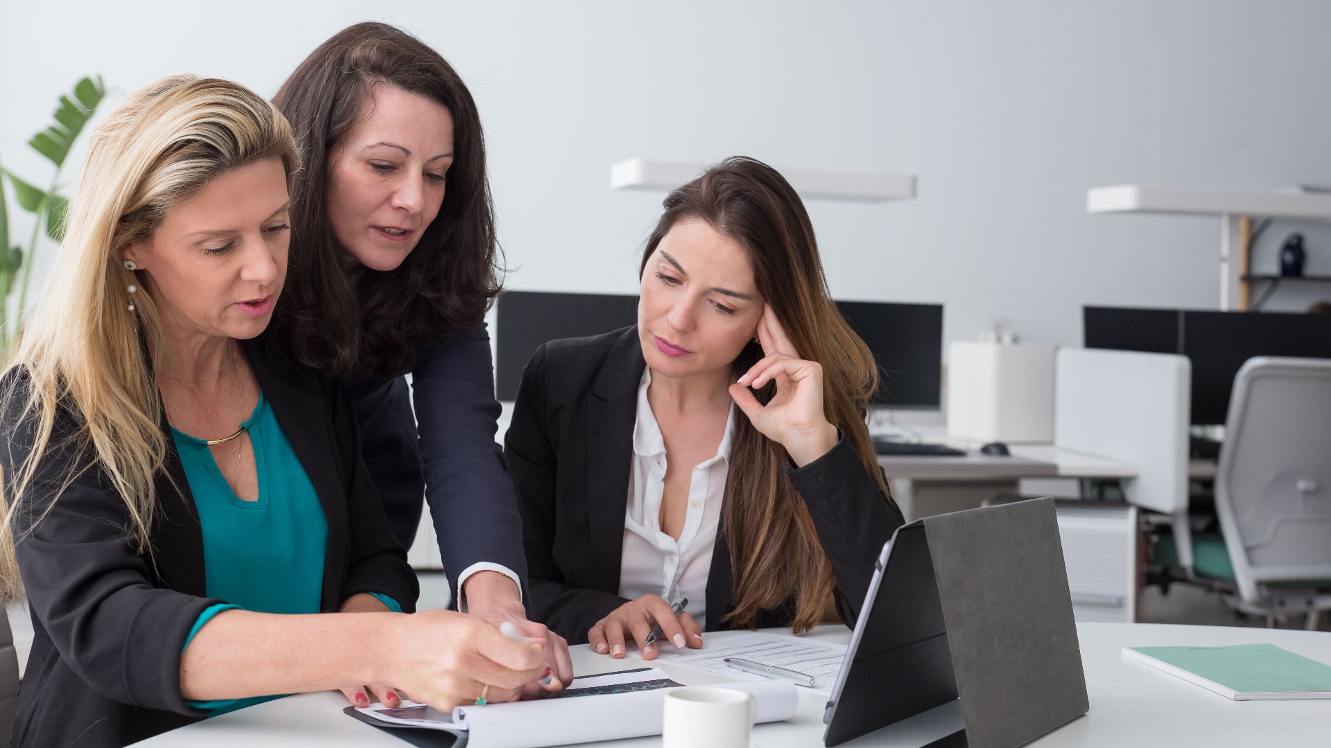 Three businesswomen collaborate on documents in a modern office setting.