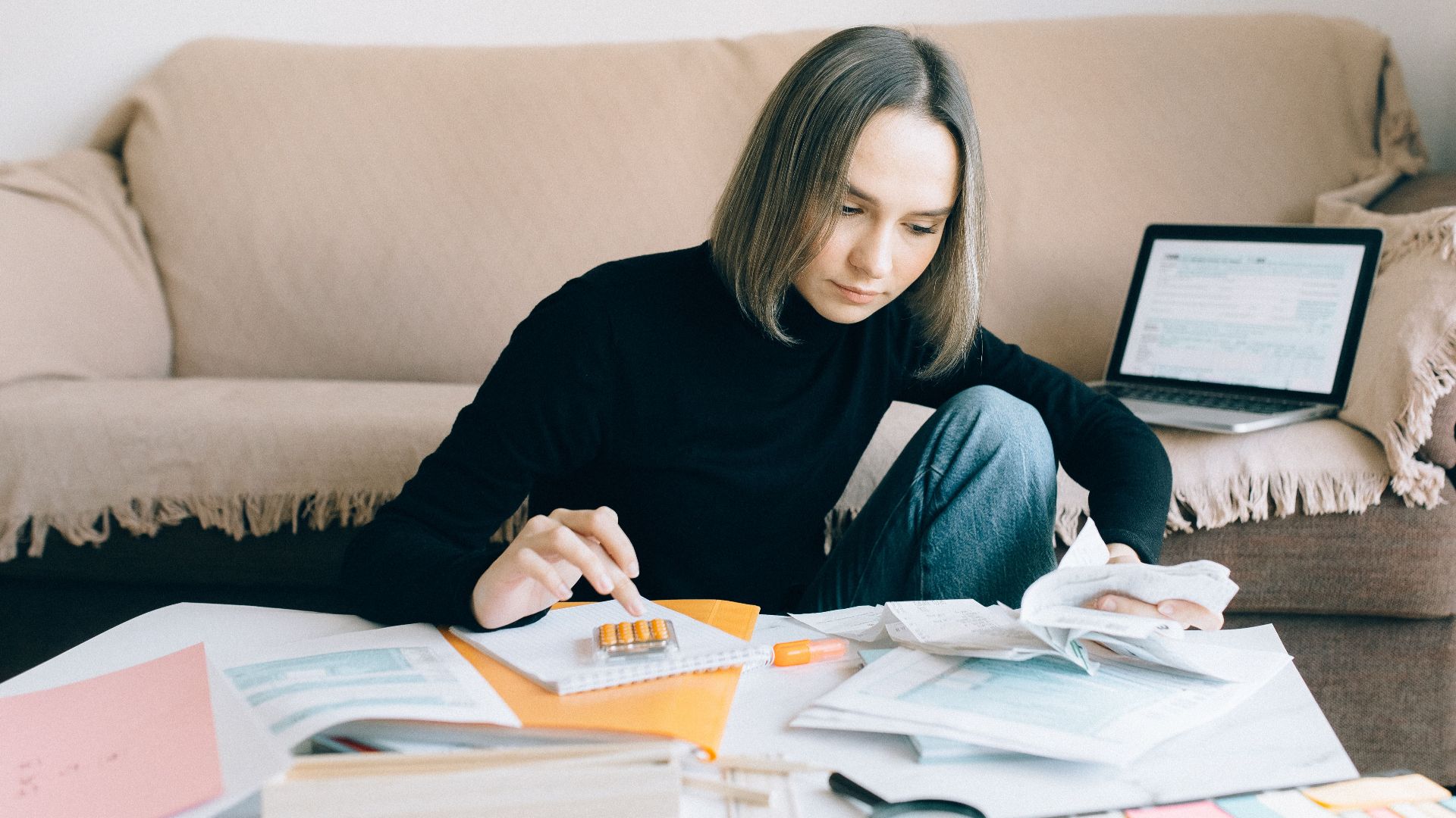 Young woman handling financial tasks with papers and laptop in cozy living room.
