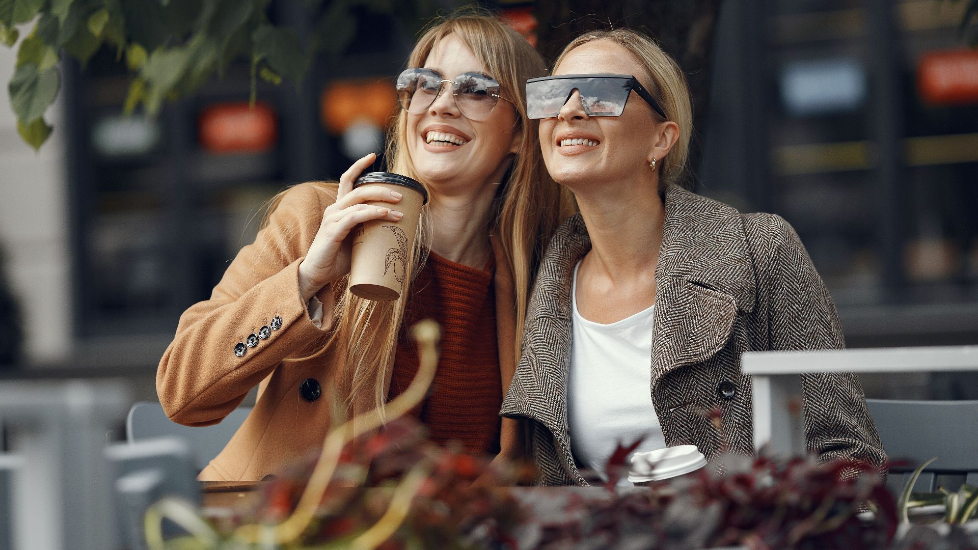 Two women sitting outdoors, smiling and enjoying coffee, wearing sunglasses.